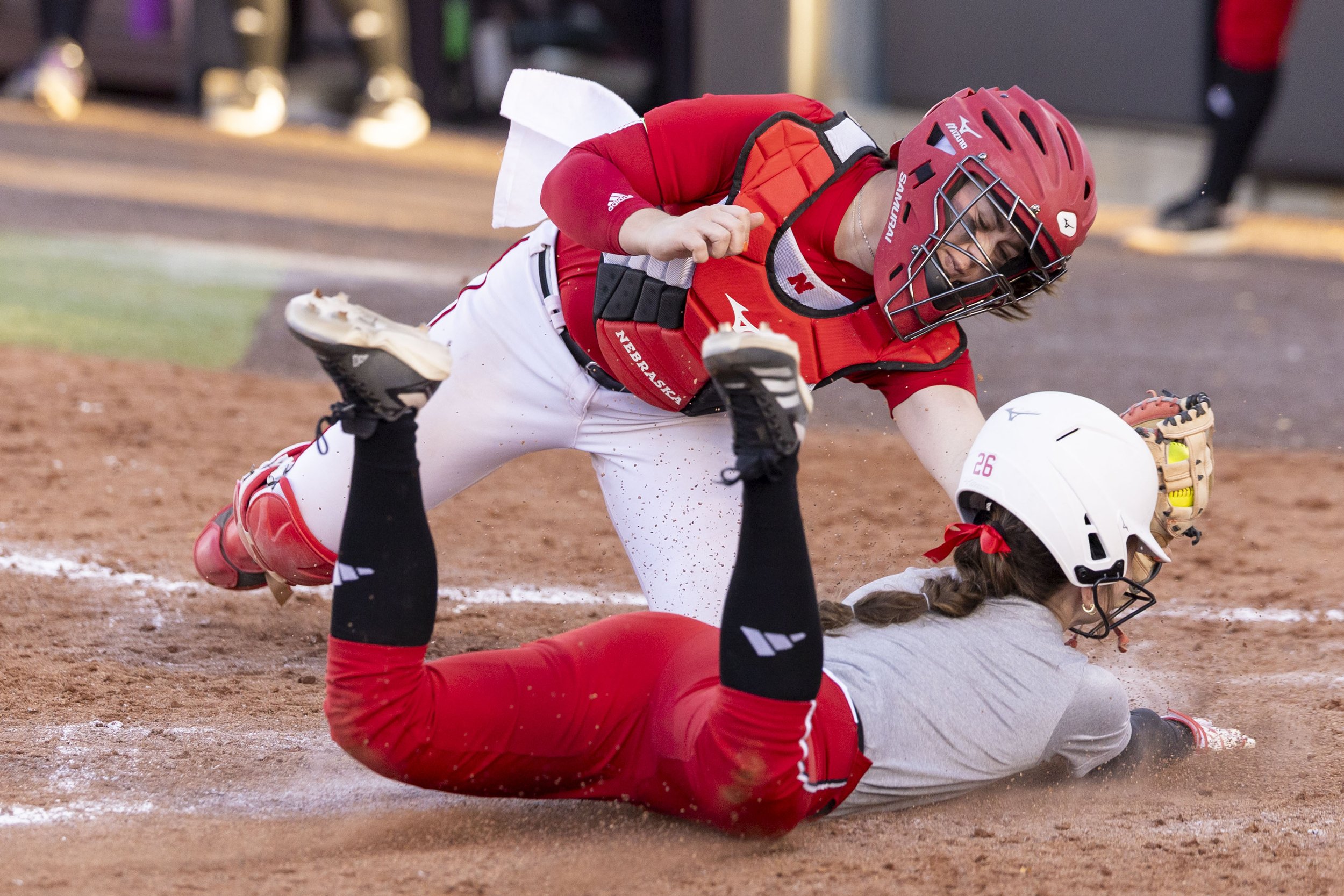 Two female softball players in red and gray uniforms collide on the dirt infield. One player, wearing a catcher's gear including a helmet, chest protector, and leg guards, is falling forward while the other, on the ground, is trying to reach for the 