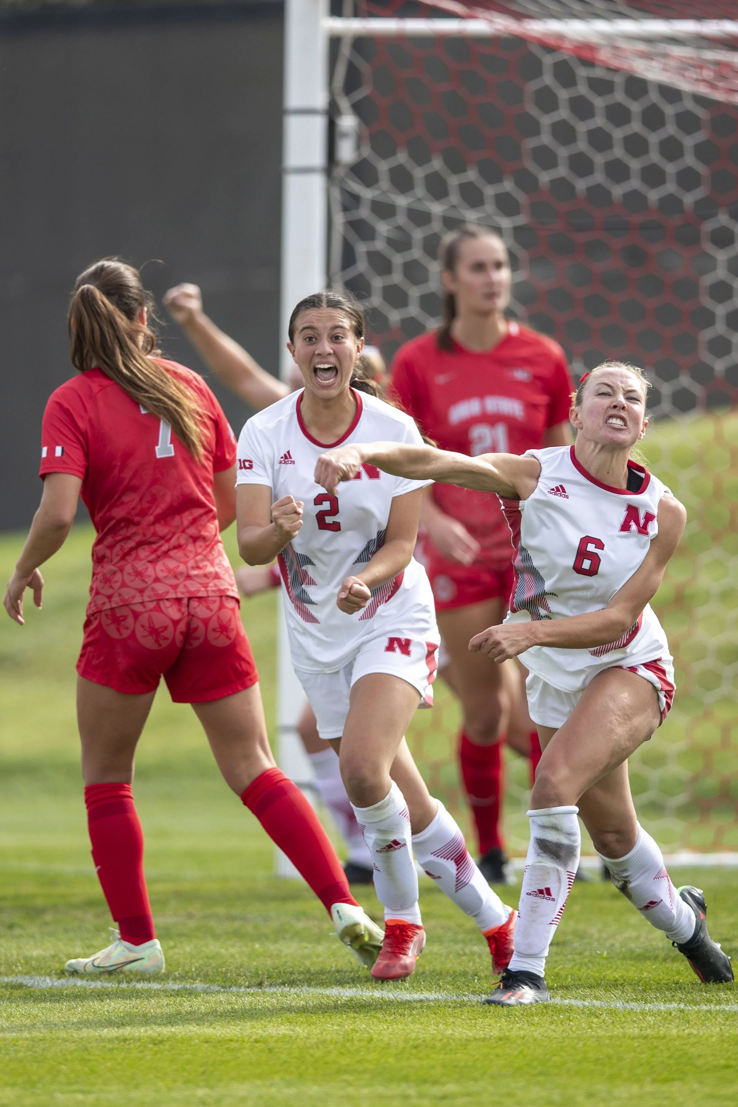 Women’s soccer players celebrating a goal on the field, wearing white and red jerseys with the letter N, in front of a goal net.