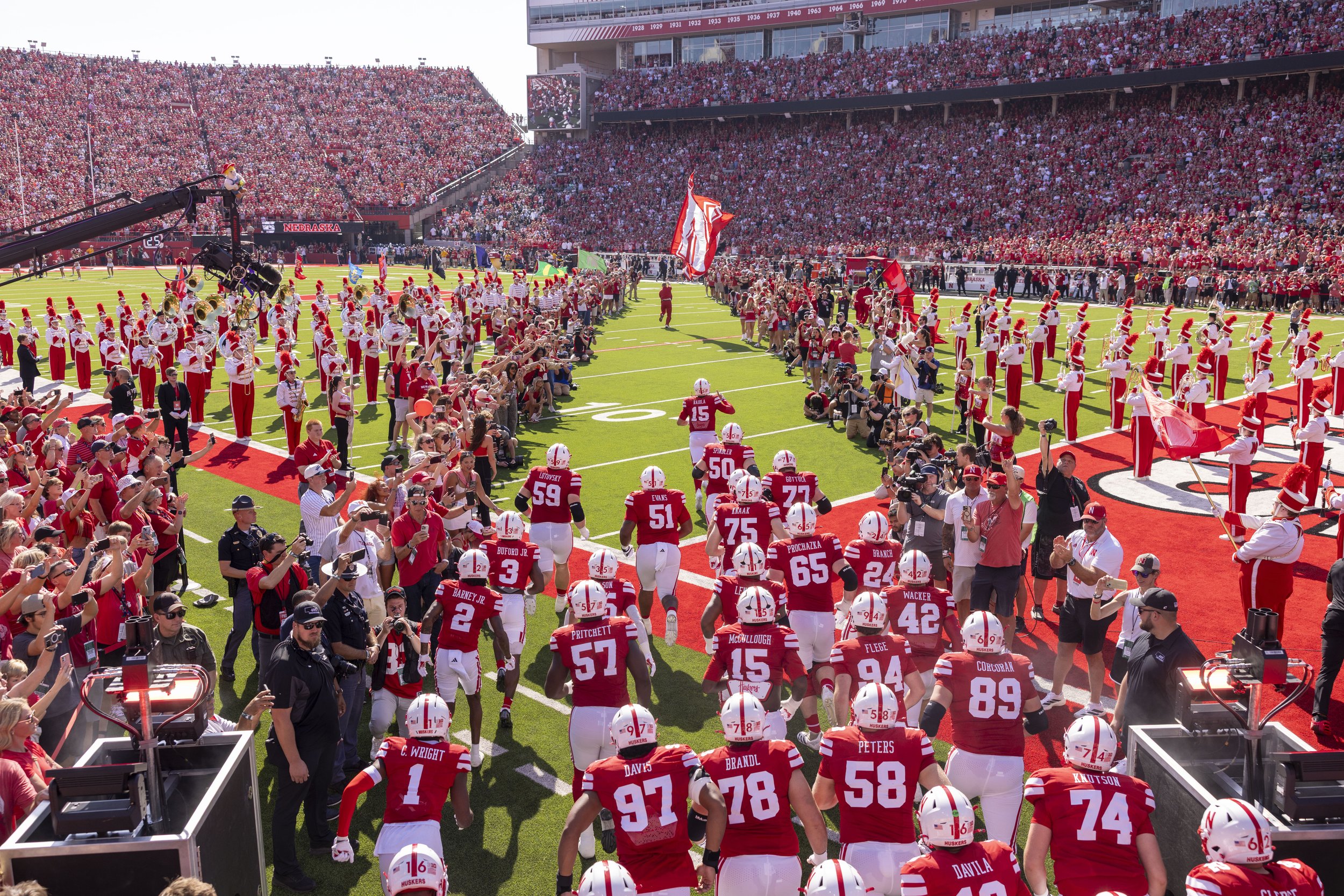 Nebraska Cornhuskers football players entering the field through a sea of fans, with a marching band on the right side and a crowd filling the stadium in the background.
