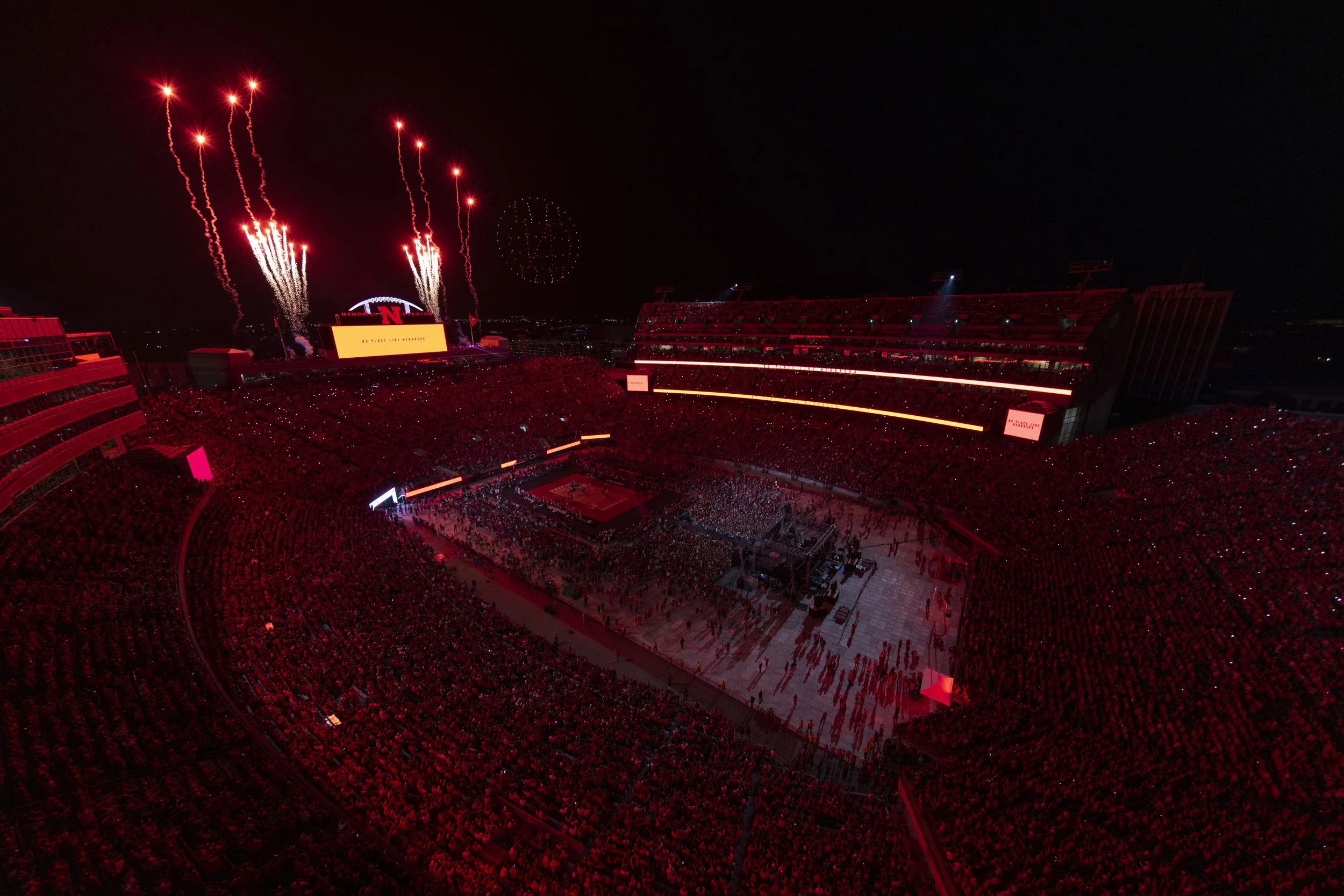 A nighttime scene of a packed sports stadium with fireworks in the sky and a large crowd on the field, illuminated by red lighting.
