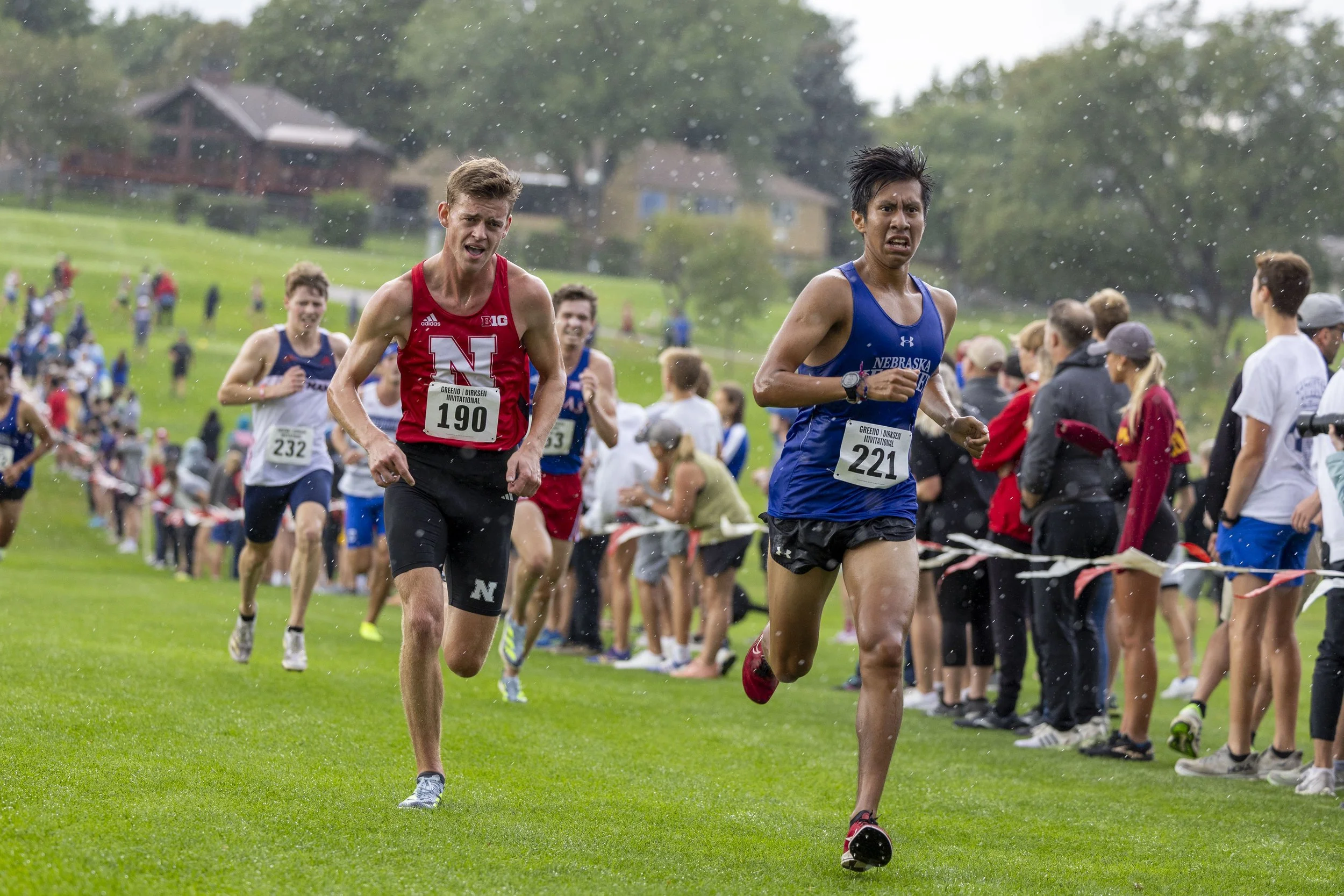 Runners competing in a cross-country race on a grassy field on a rainy day, with spectators watching along the sidelines.