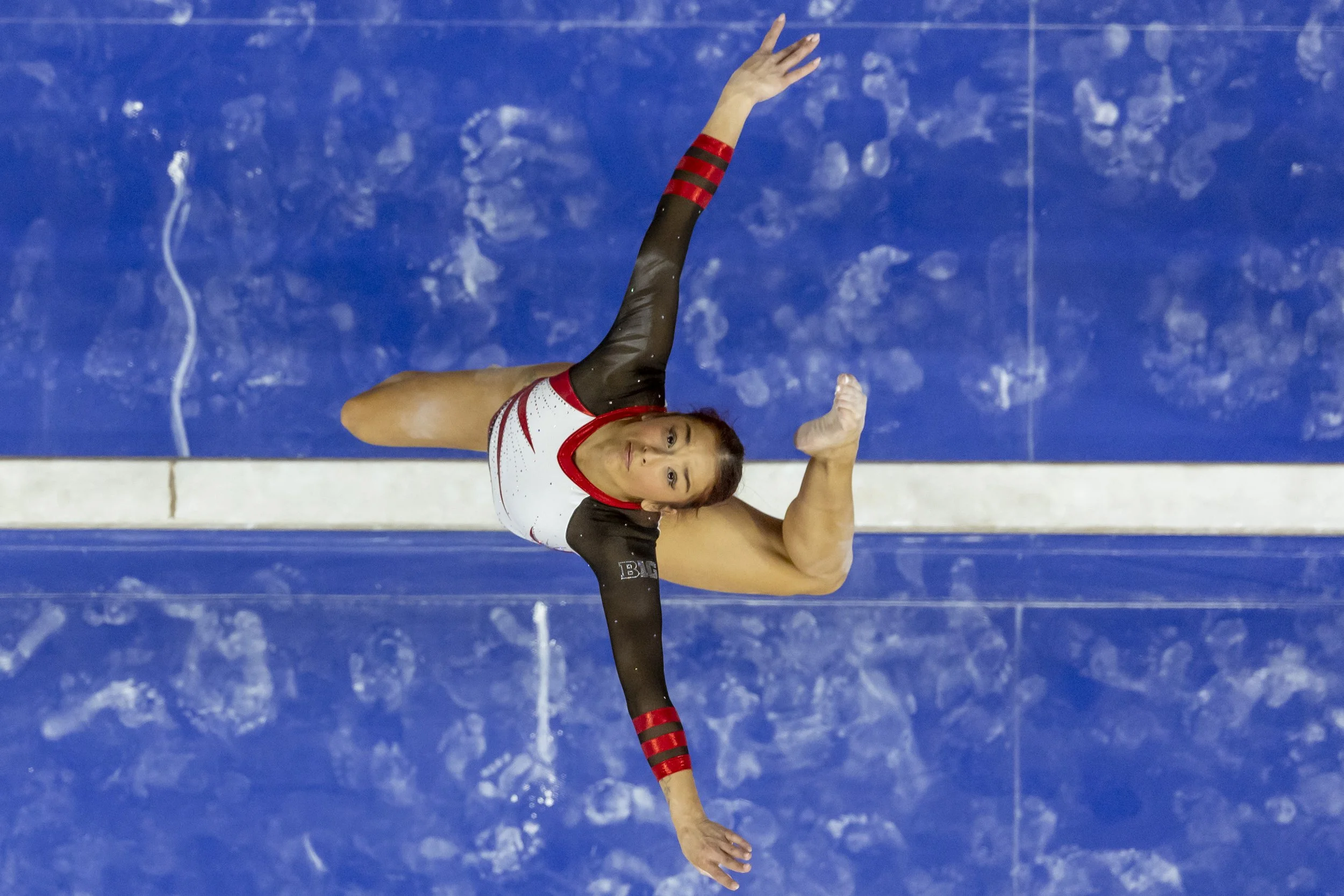 Female gymnast performing a floor routine on a blue mat, wearing a black, white, and red leotard, viewed from above.