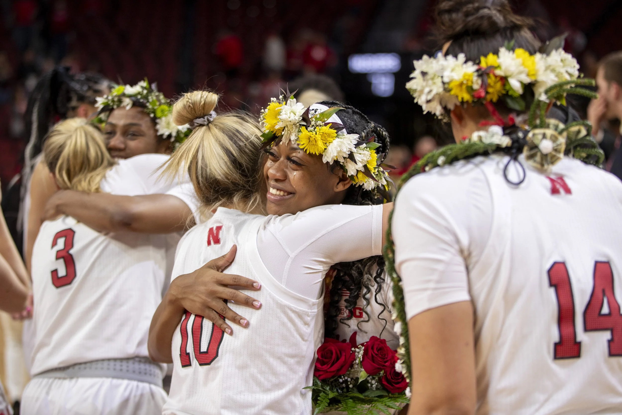 A group of female athletes wearing white jerseys with red numbers and floral crowns are hugging and smiling during a celebration. One woman in the center is holding a bouquet of red roses.