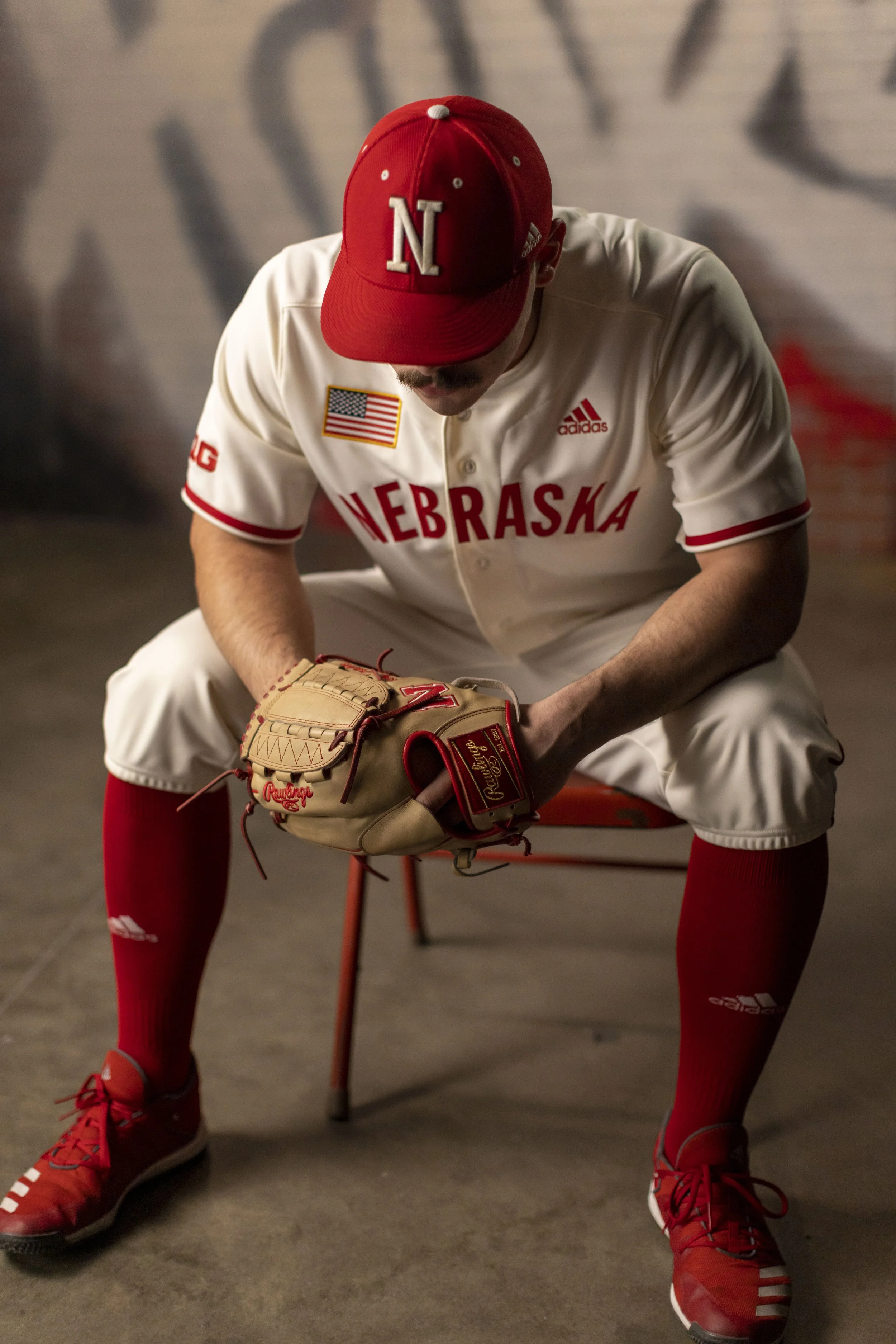 A man dressed in a Nebraska baseball uniform with a red cap, white jersey, and red socks, sitting on a red chair with a baseball glove on his hand, in front of a graffiti wall.