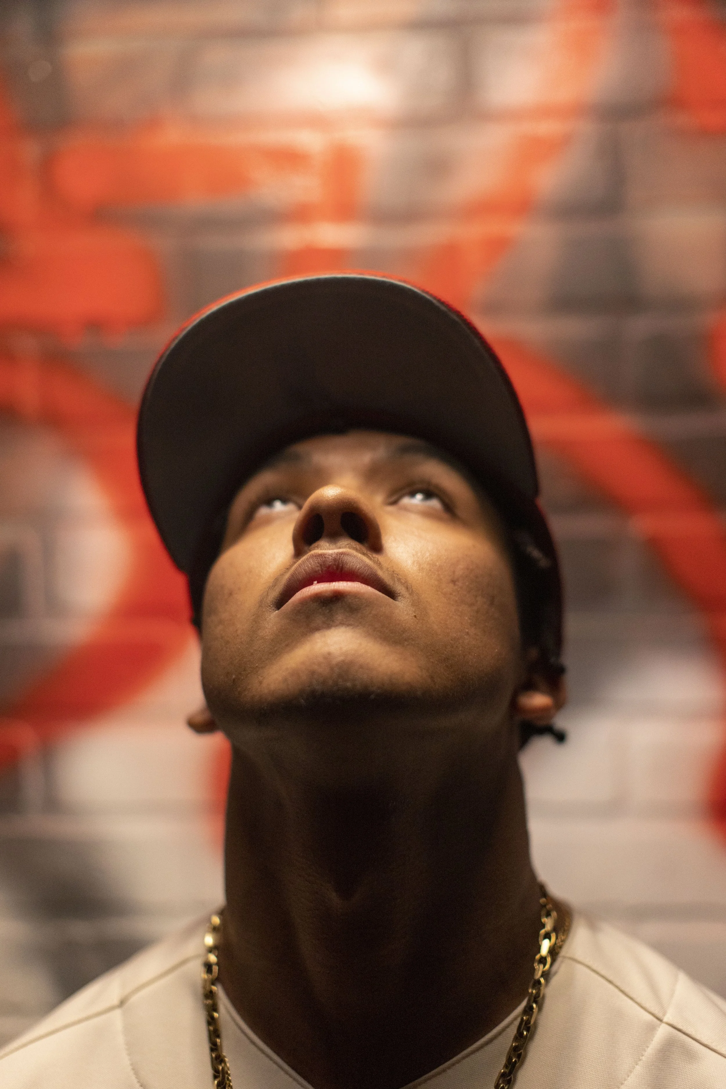 Close-up of a young man wearing a black baseball cap, looking upward against a graffiti-covered brick wall background.