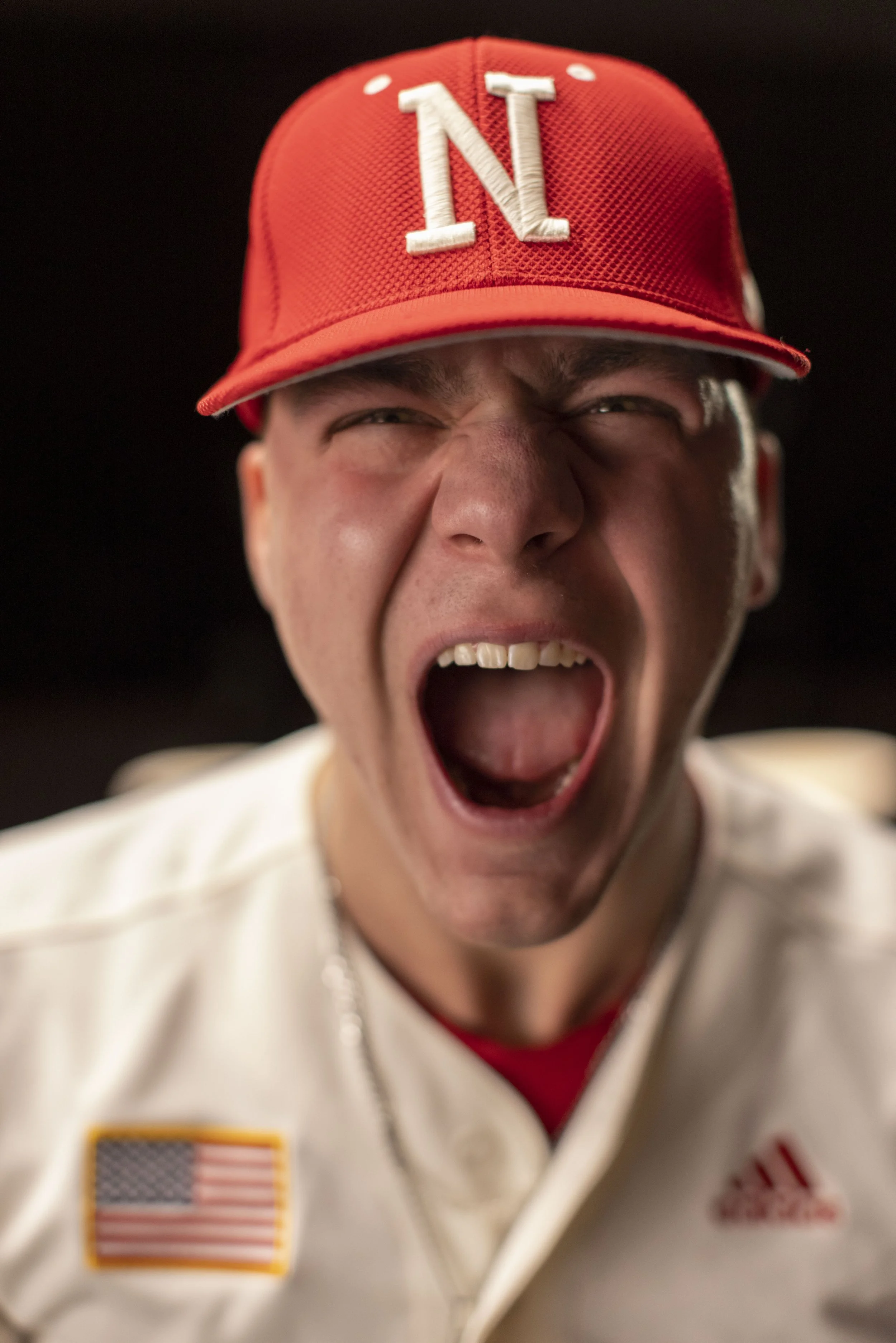 A young man wearing a red baseball cap with the letter 'N' and a white jacket with an American flag patch and an Adidas logo, yelling with his mouth open and eyes squinted.