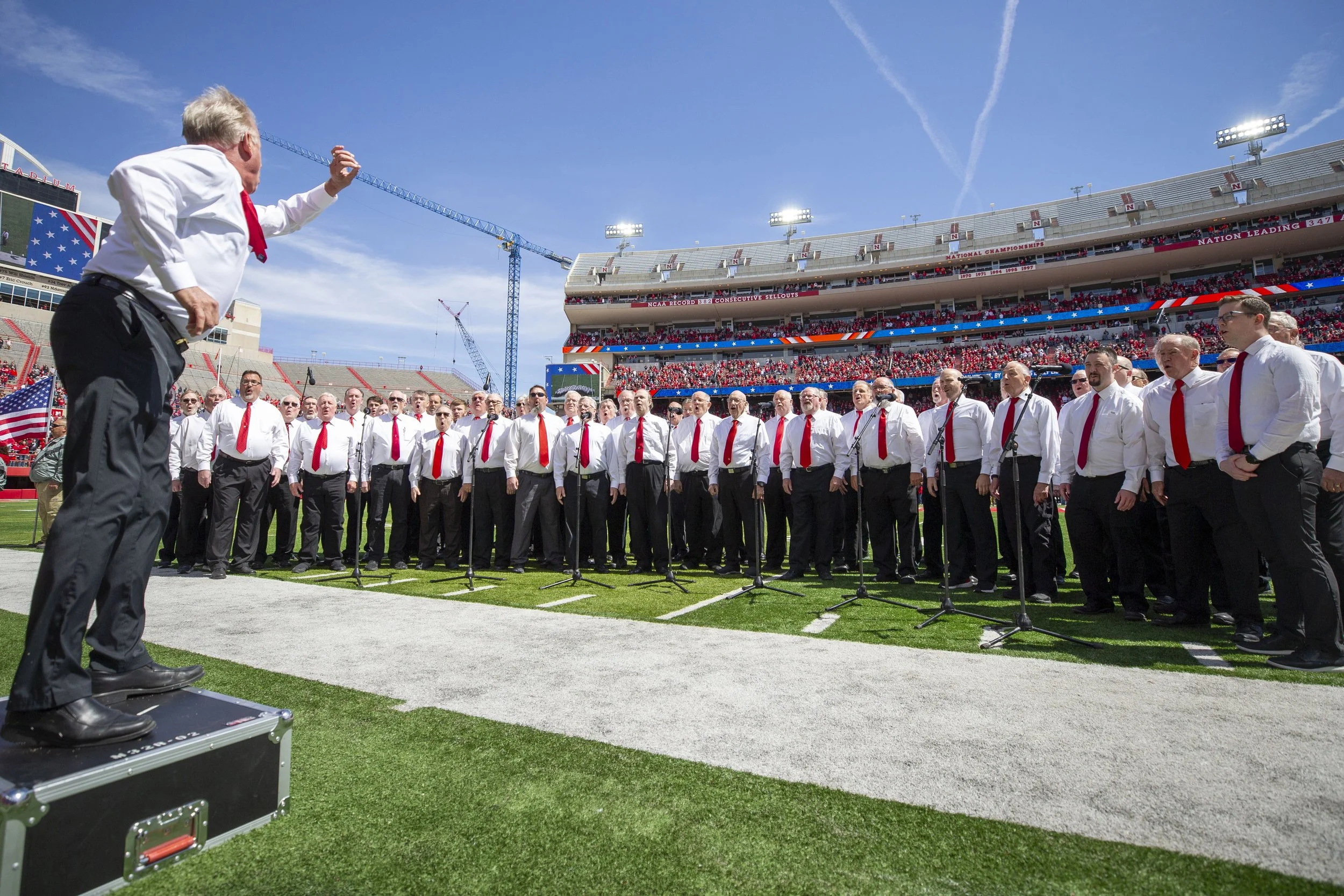 A choir of men wearing white shirts and red ties performing on a football field, directed by a man conducting from a platform, with a large stadium and crowd in the background on a sunny day.