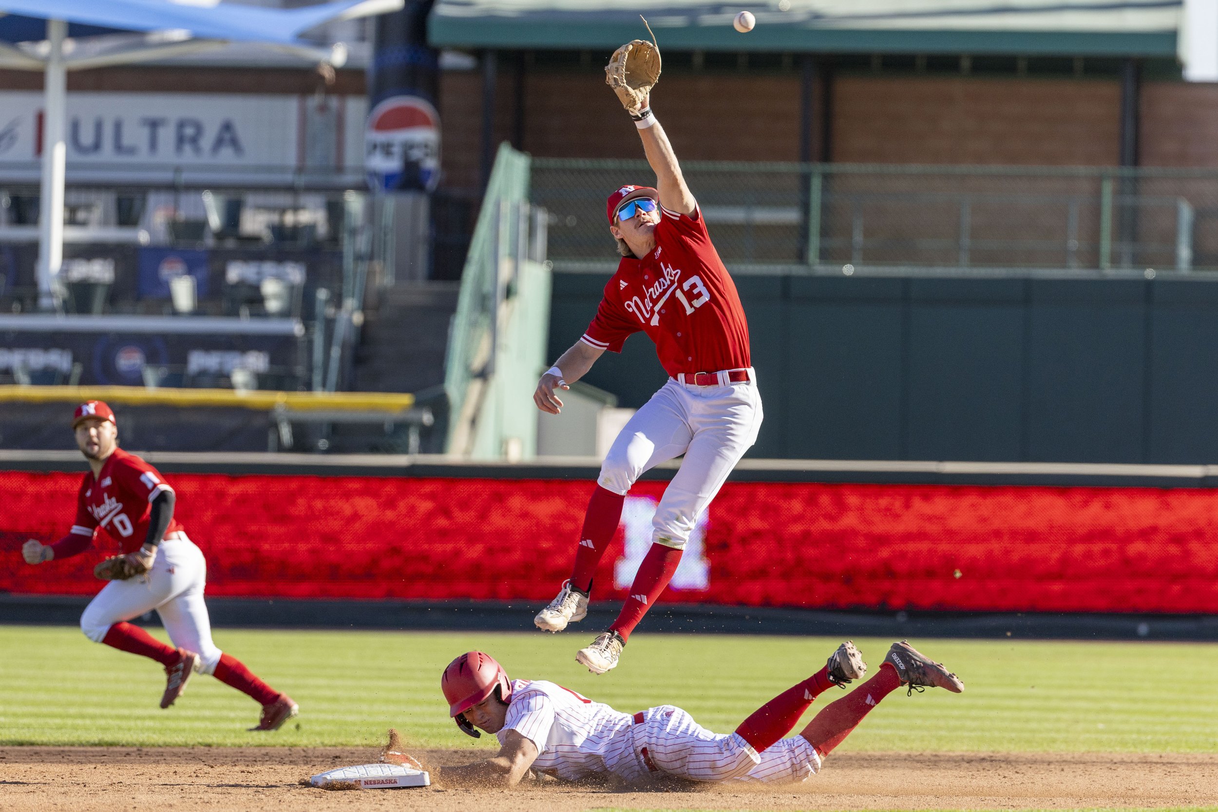 A baseball player in a red jersey and white pants is leaping in the air to catch a ball with his glove, while another player slides into the base on the dirt field. Two other players are visible in the background, and the scene is set on a sunny base