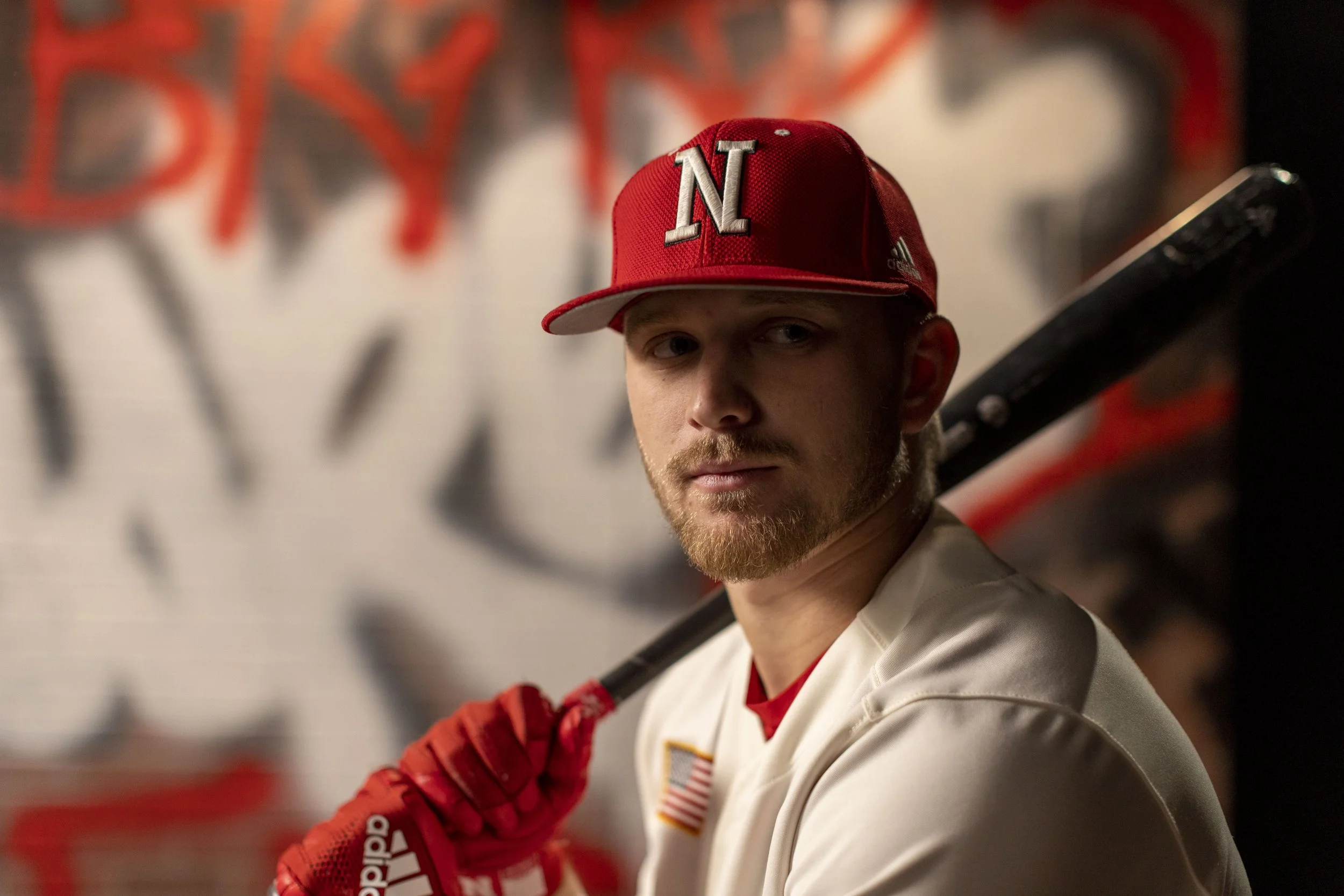A young man in a white baseball uniform with an American flag patch, wearing a red baseball cap with a white N, holding a black bat over his shoulder, standing in front of a graffiti-style wall.