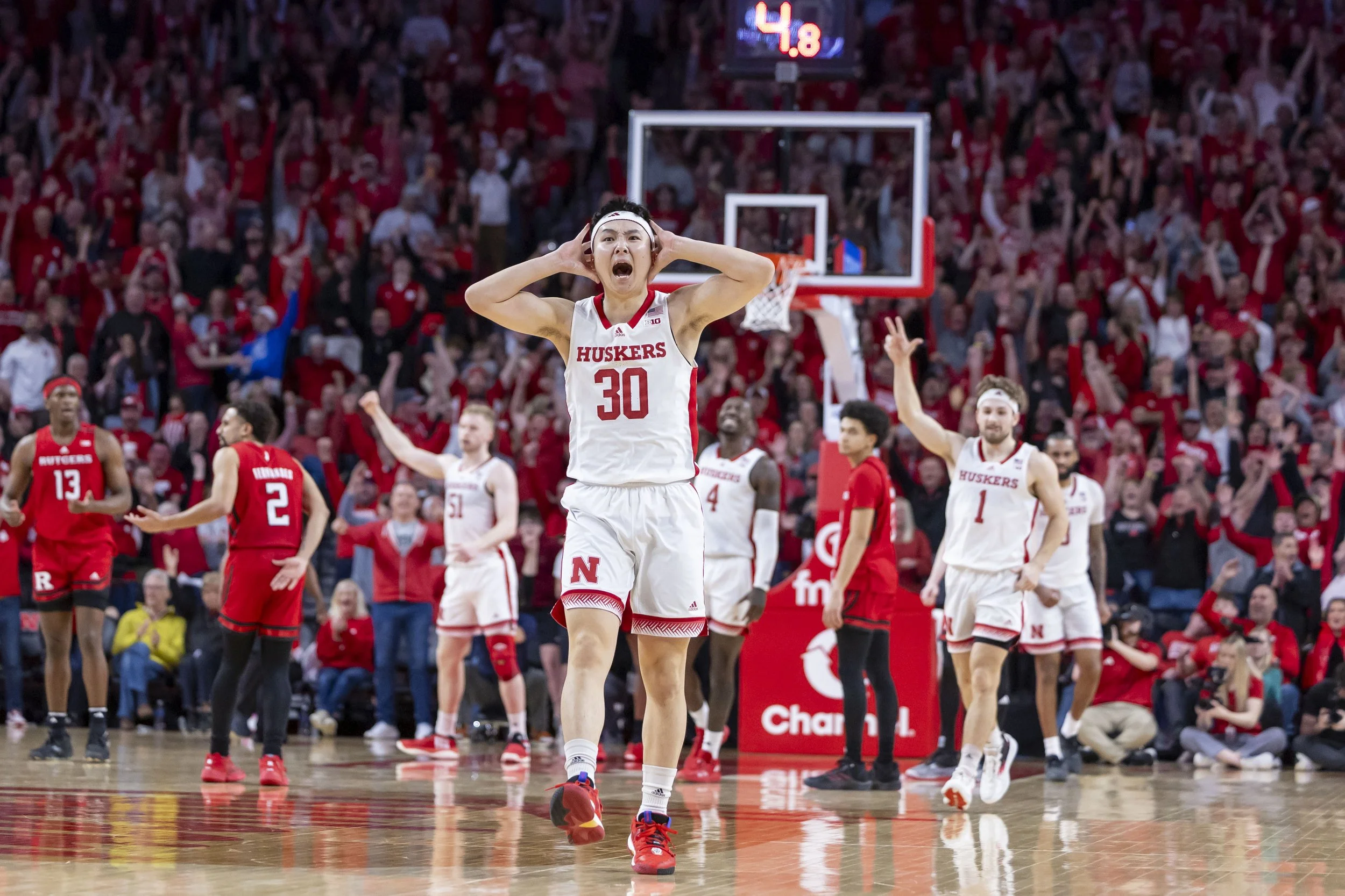 A women's college basketball game showing players from Nebraska Huskers and Wisconsin Badgers, with Nebraska player number 30 in the foreground holding her head with a surprised or upset expression, and a crowded arena filled with fans in red and whi