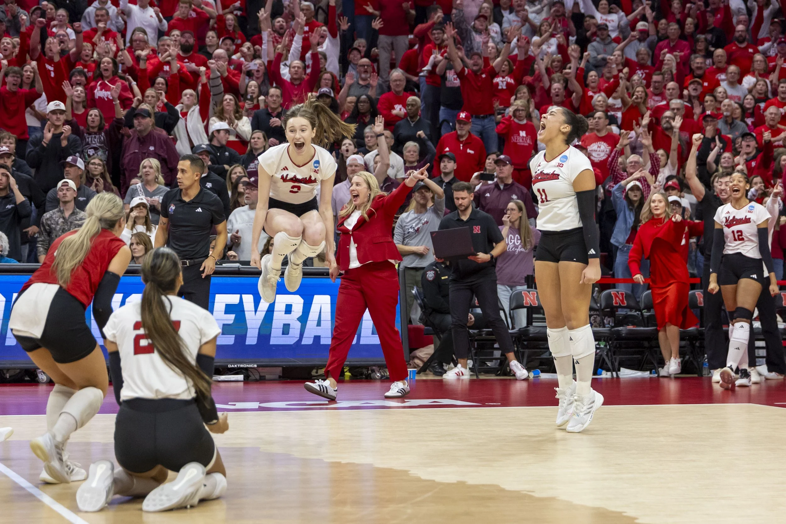 A volleyball player wearing jersey number 3 is jumping in celebration while her teammates and crowd cheer around her on the court.