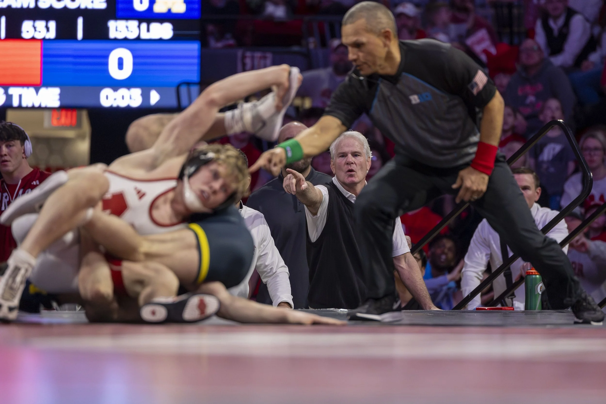 Two wrestling athletes on the mat during a match, with a referee intervening and pointing to a call, in front of a crowd and scoreboard in the background.