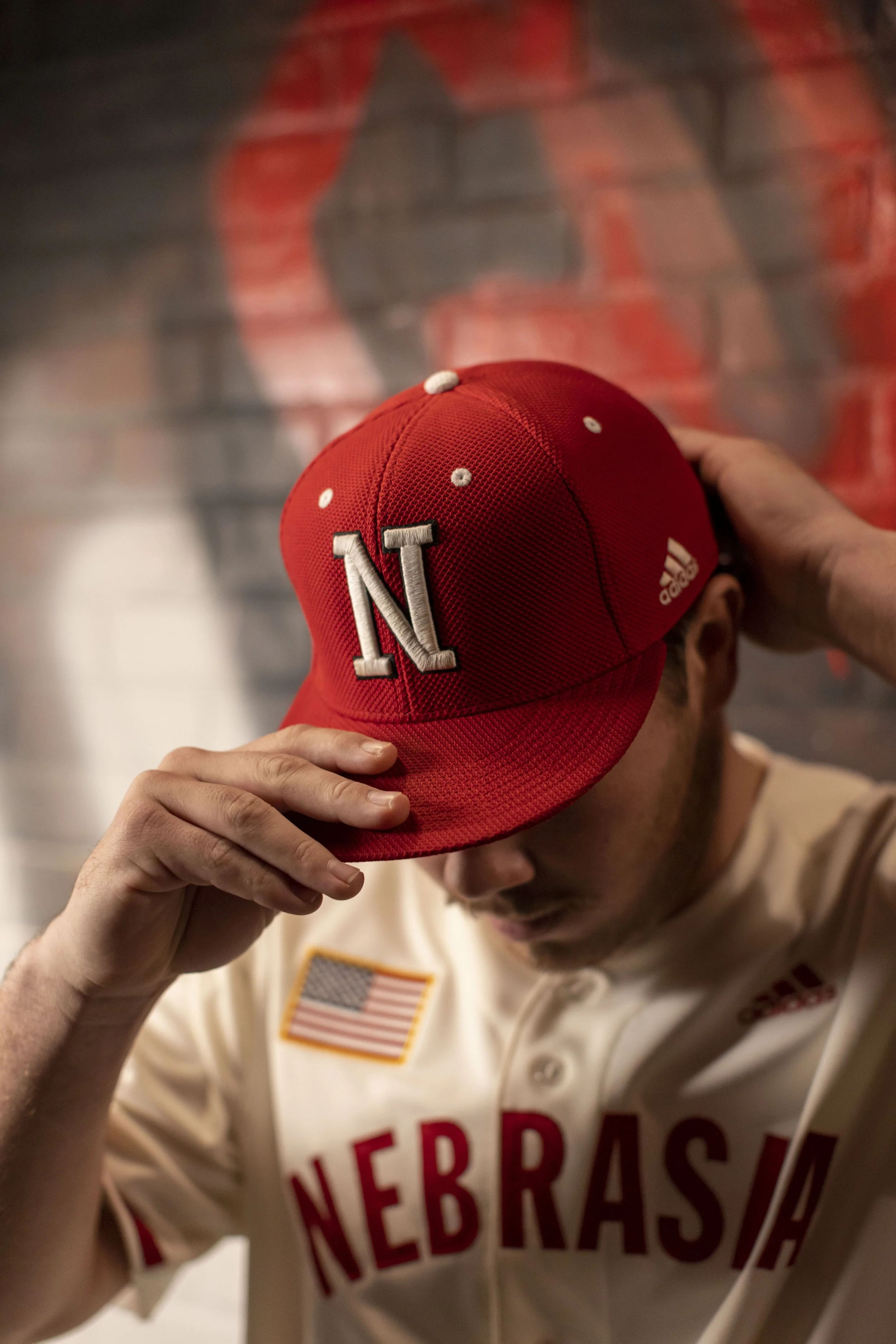 A man wearing a Nebraska Cornhuskers baseball jersey adjusts a red Nebraska baseball cap, with a brick wall in the background.