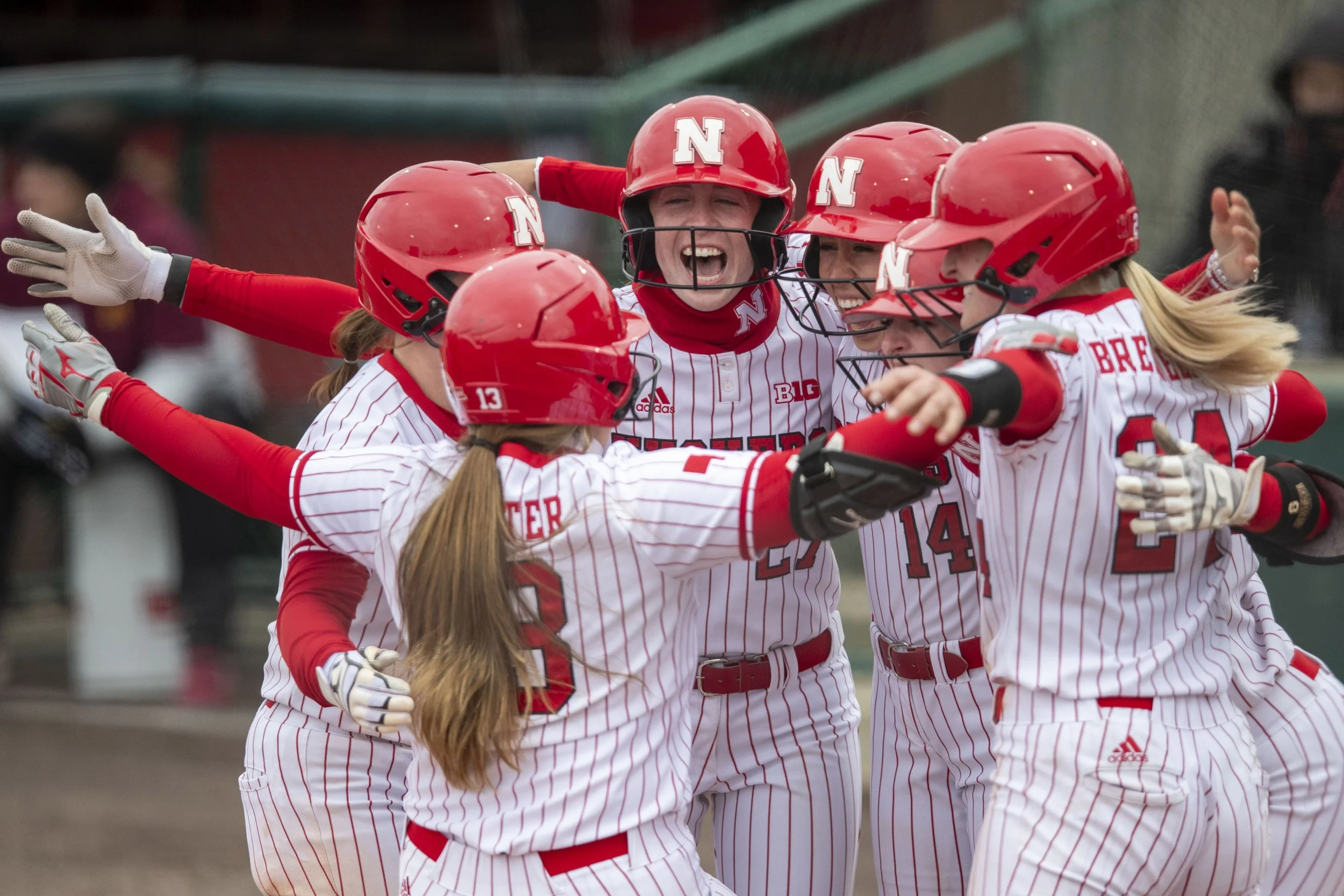 A group of female softball players in red and white striped uniforms with red helmets celebrating together on the field.