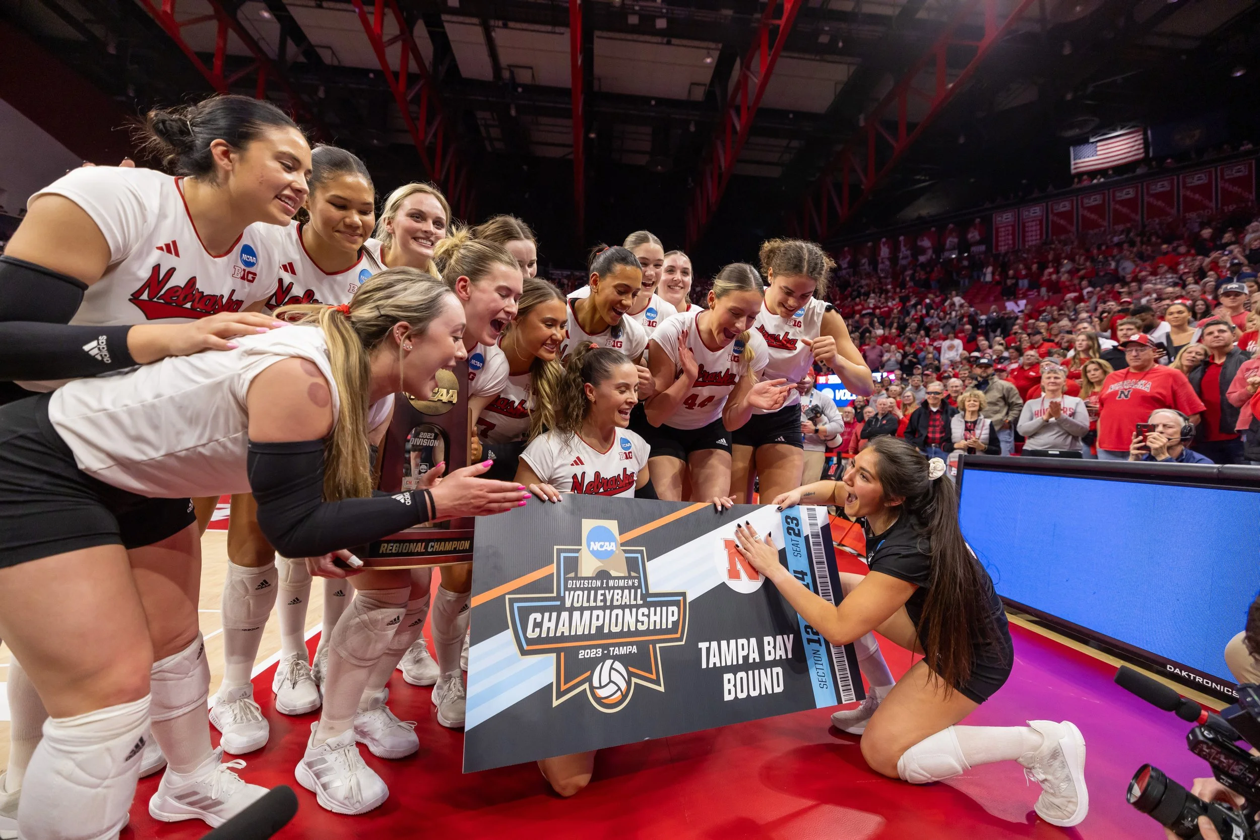 A group of female volleyball players in white uniforms celebrating after winning a championship, holding a large trophy and a sign that reads 'NCAA Division I Women's Volleyball Championship, Tampa Bay Bound 2023.'