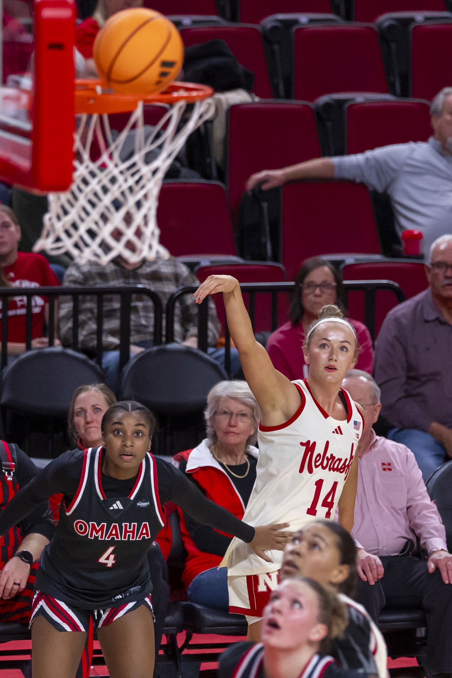A women's college basketball game with a player from Nebraska shooting and a player from Omaha trying to block, while spectators watch.