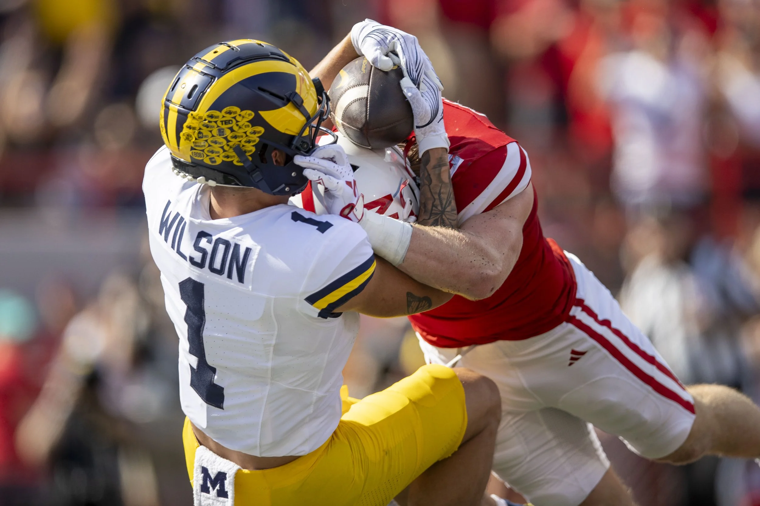 A football player in a red and white uniform is tackled by a player in a white and yellow uniform with a Michigan helmet during a game.