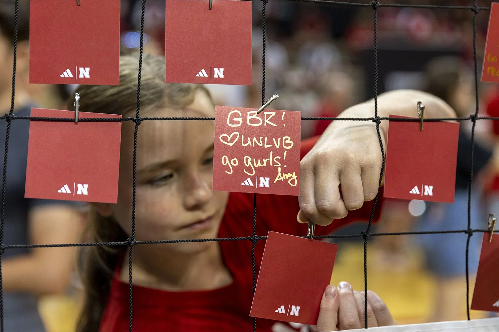A young woman attaches red note cards to a black wire grid. One note card has a message written in yellow marker: "GBR! ♥ UNLVB go gurs! Amy," with the University of Nebraska logo in the bottom corner.