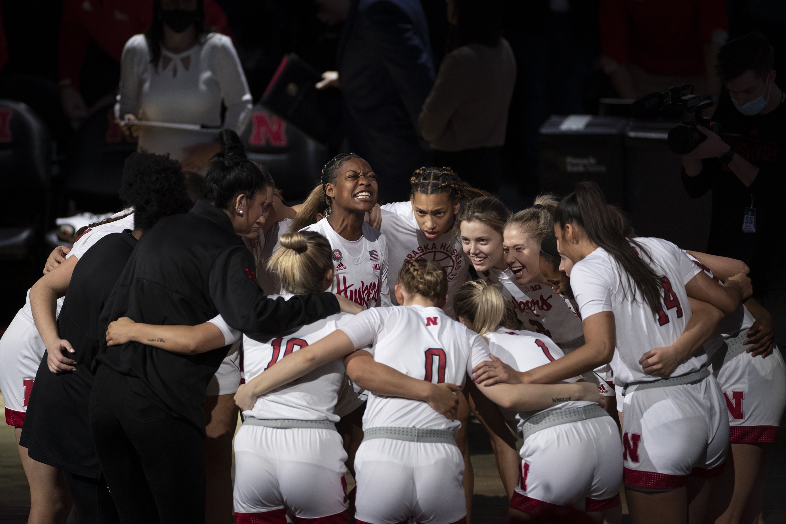 Nebraska women's volleyball team huddled together during a moment of prayer or motivation on the court, wearing white uniforms.