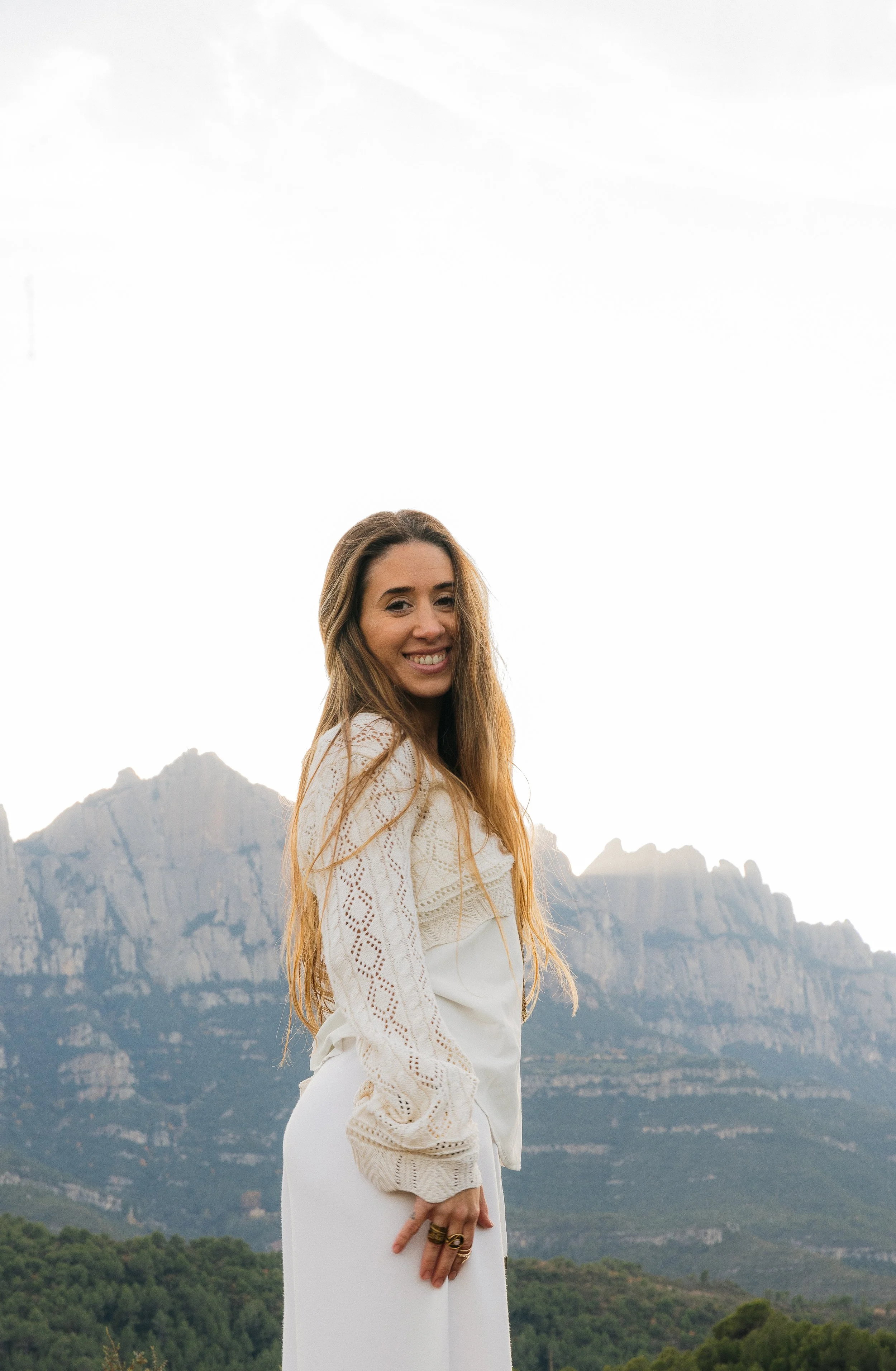 A woman with long brown hair smiling and looking at the camera, standing outdoors in front of mountains and a cloudy sky.