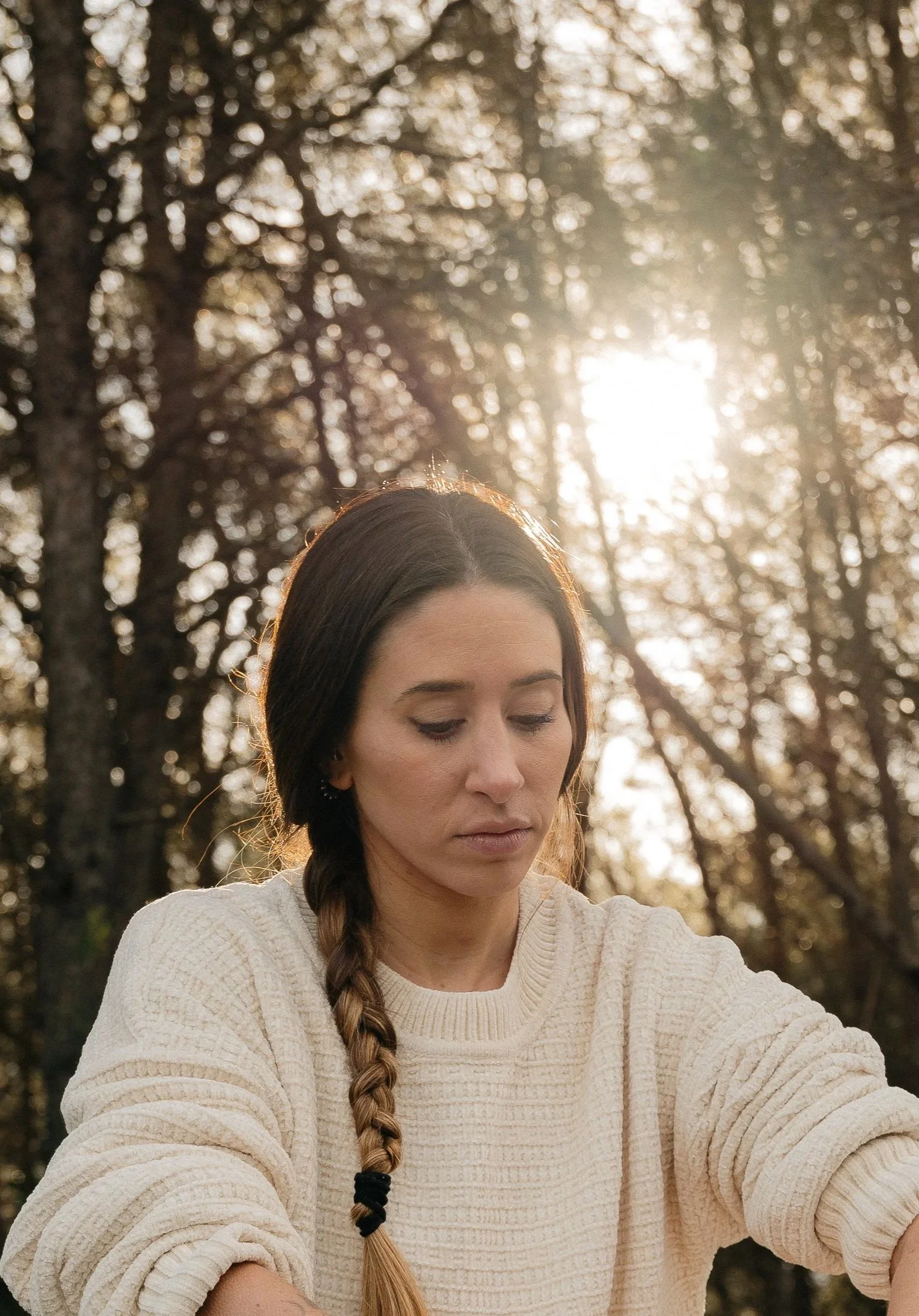 A woman with long brown hair in a braid, wearing a cream-colored sweater, looking down thoughtfully outdoors during sunset or sunrise with trees in the background.