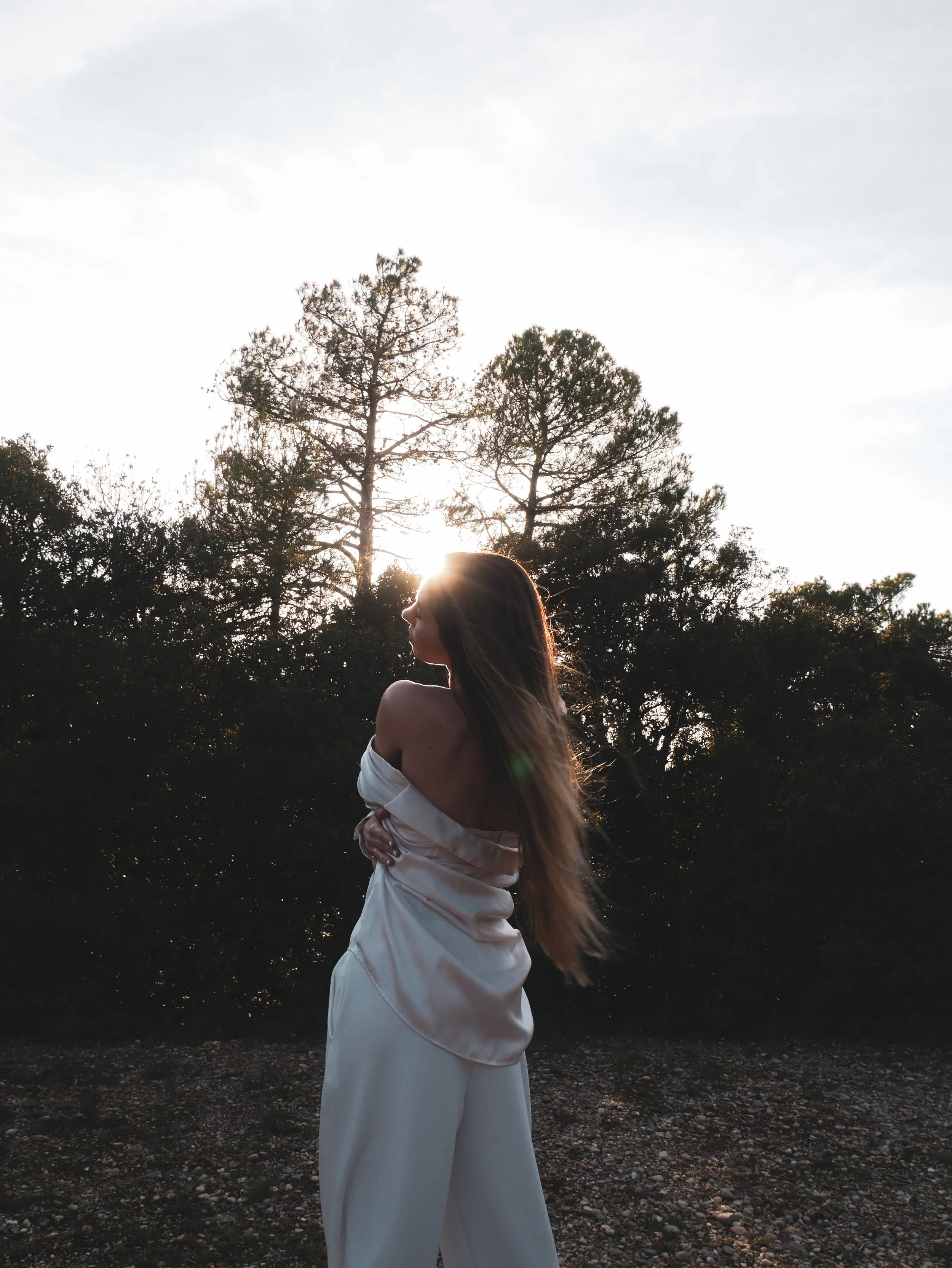 A woman in white clothing standing outdoors, with her arms crossed and her eyes closed, during sunset with trees in the background.