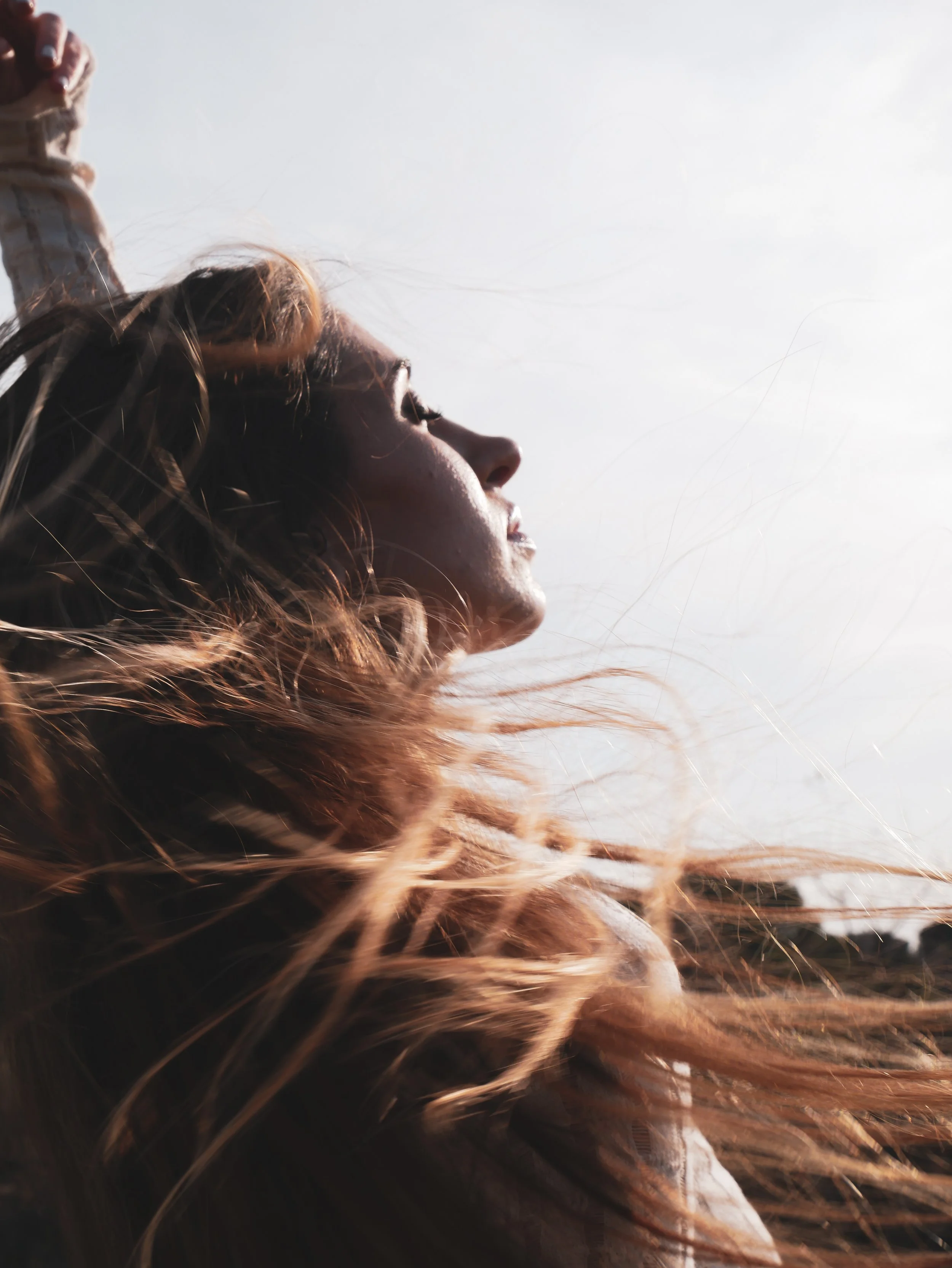 Profile of a woman with long, flowing hair blowing in the wind against a bright sky.