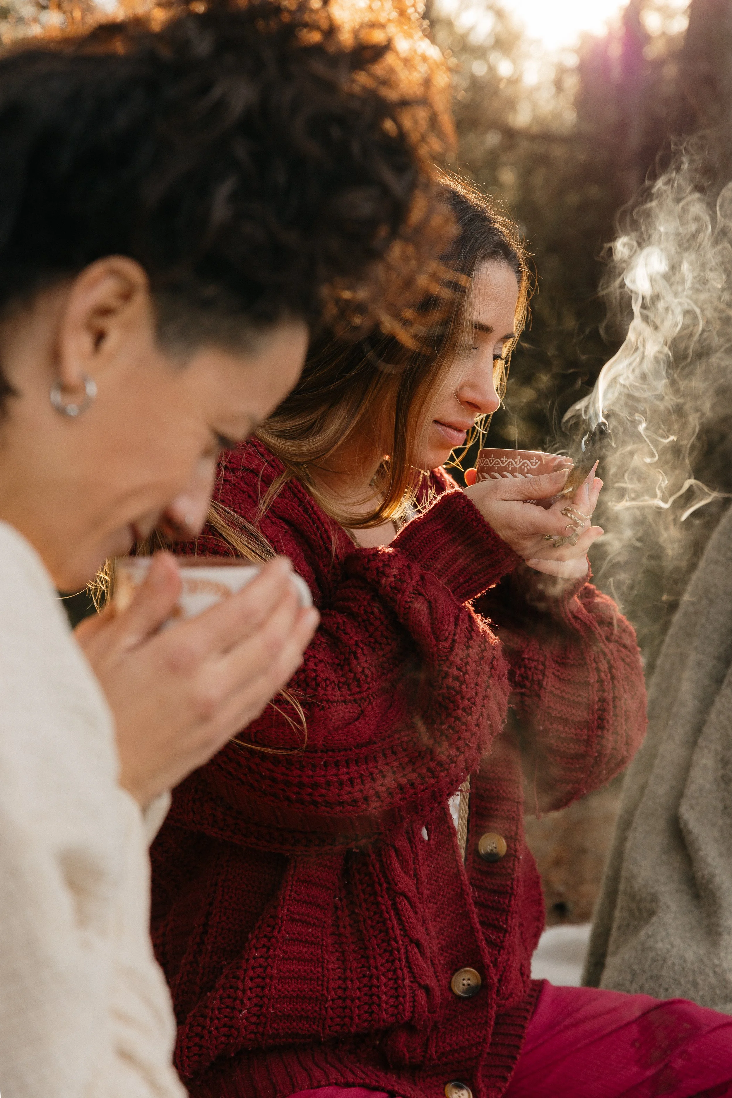 Two women in cozy sweaters enjoying hot beverages outdoors at sunset, with one woman smiling and the other with eyes closed, surrounded by autumn foliage.