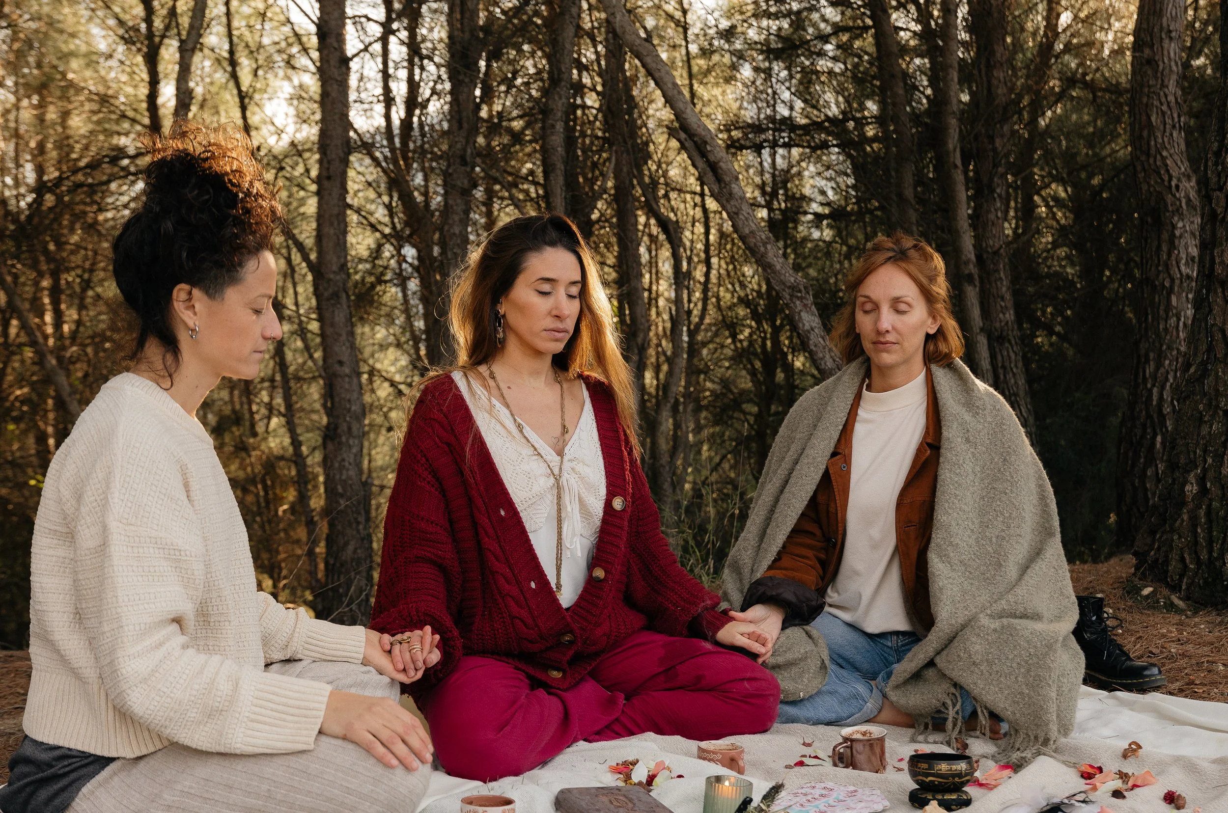 Three women sitting on a blanket in the woods with closed eyes, holding hands in a meditation session. There are cups, candles, and flowers on the blanket.
