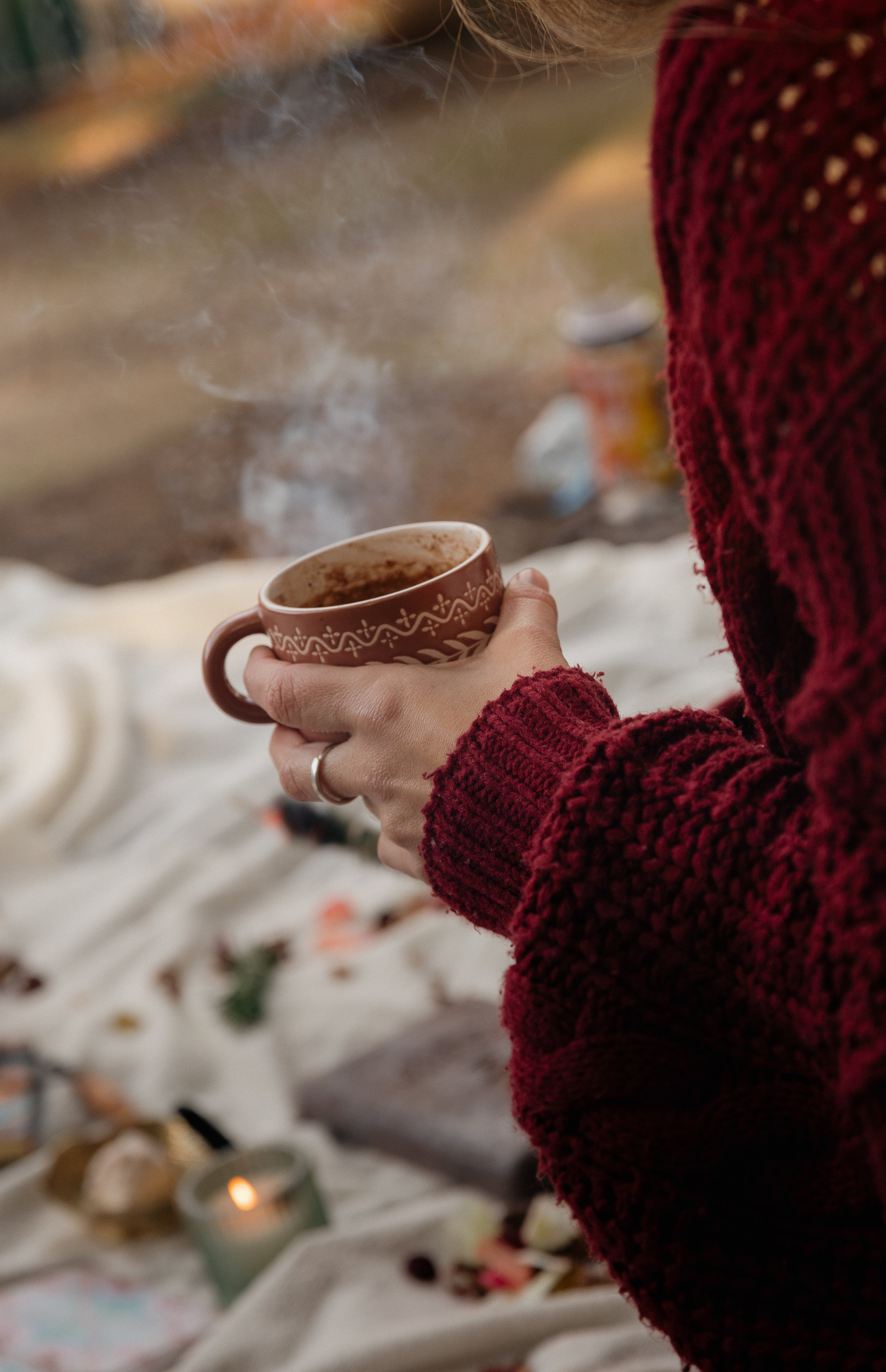 A person wearing a red knitted sweater holding a steaming hot mug with a decorative pattern outside during the daytime.