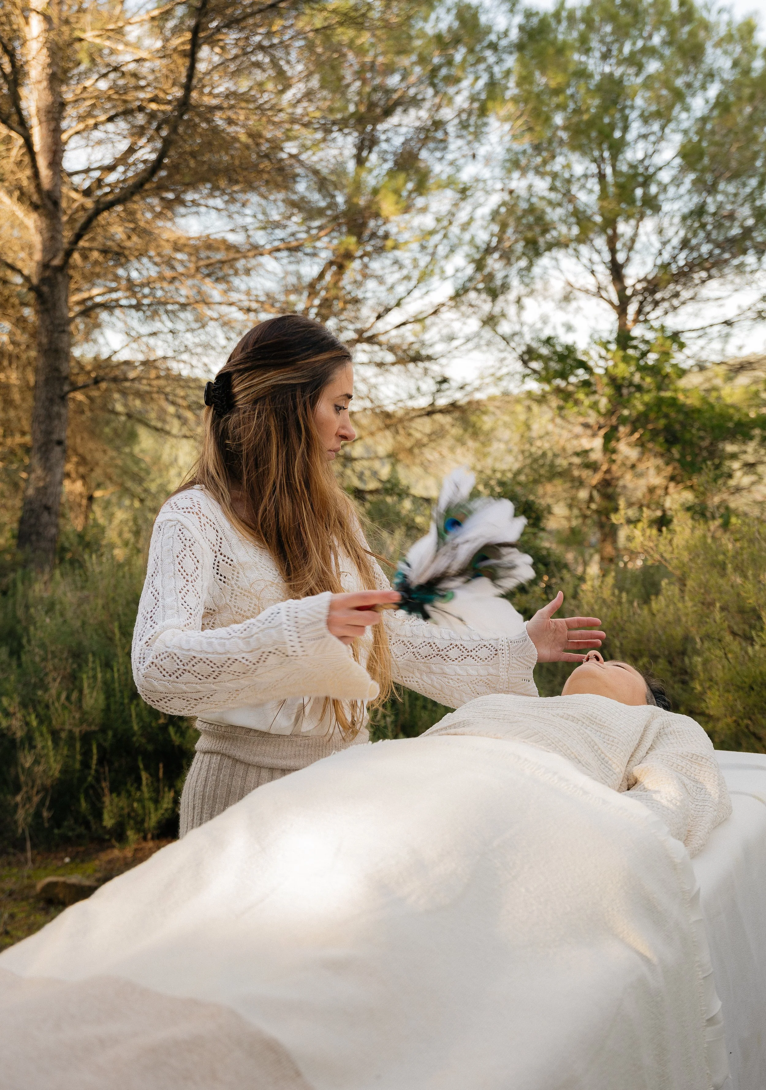Woman performing a spiritual ritual with feathers beside a person lying on a table outdoors surrounded by trees