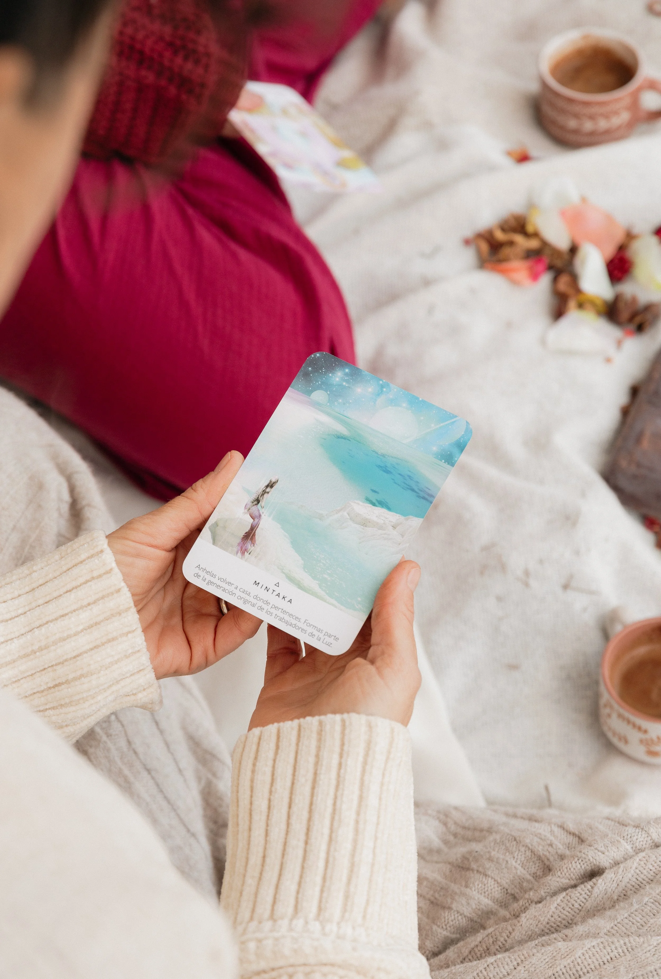 Person holding a card with a fantasy landscape of a woman on a beach, icebergs, and the sea, sitting on a blanket with coffee cups and flower petals scattered around.