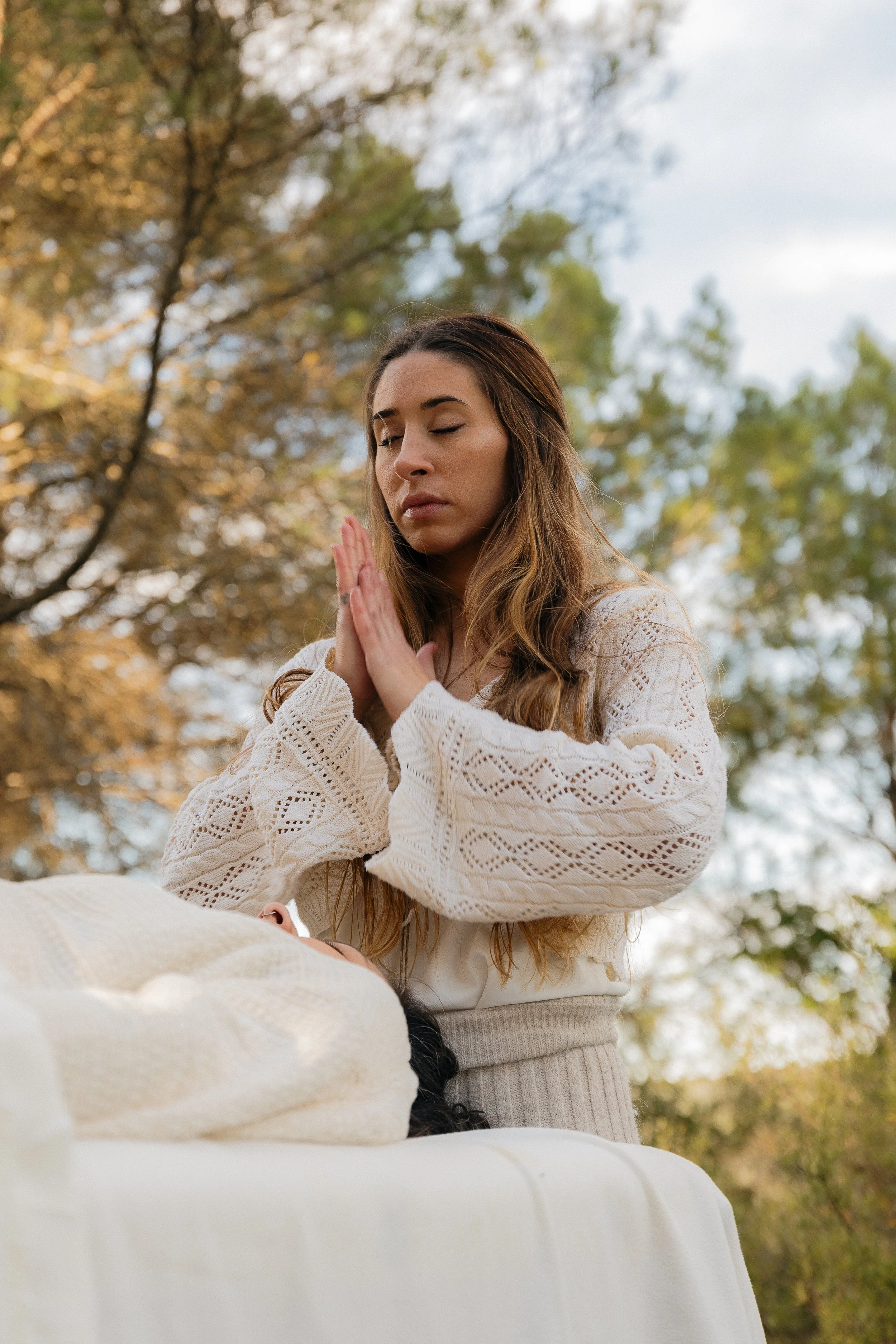 A woman with long brown hair in a white knitted sweater prays outdoors with her eyes closed, during daytime with trees in the background.