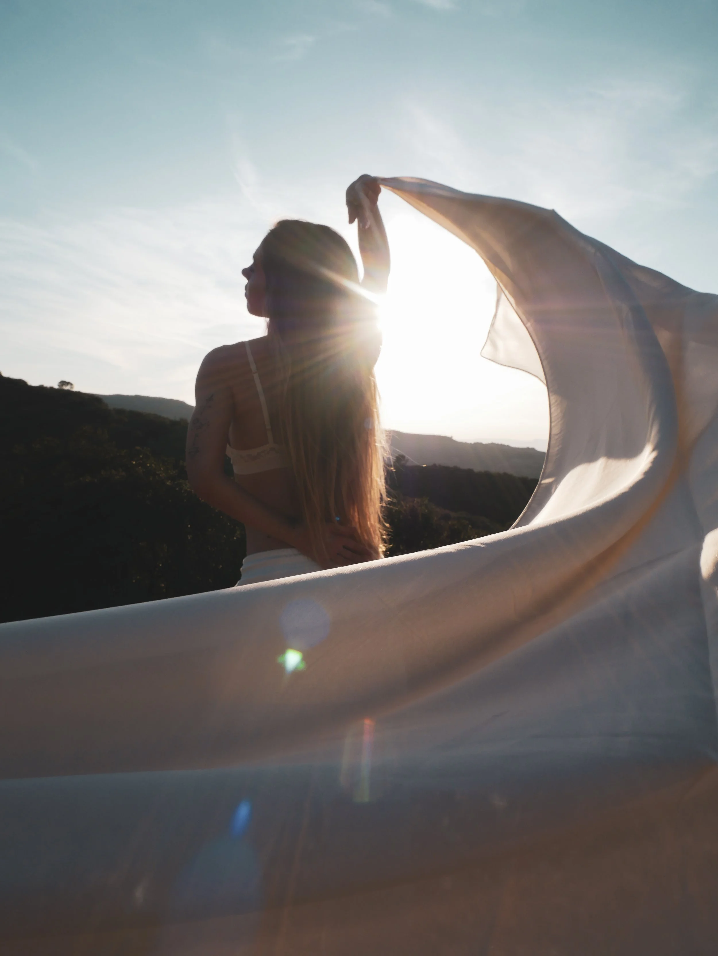 Woman holding a flowing fabric outdoors with a scenic landscape and a setting sun behind her.
