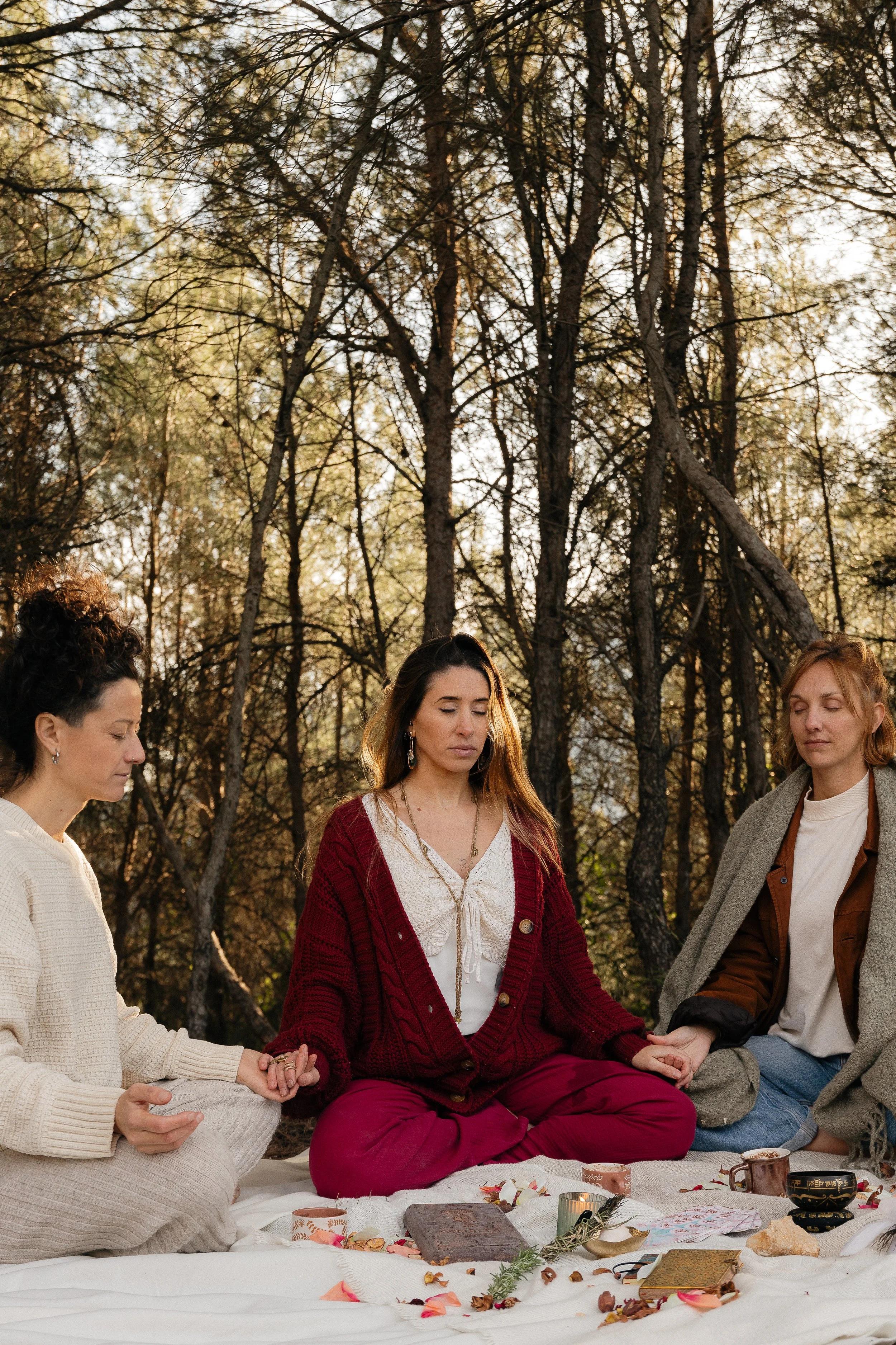 Three women sitting cross-legged and holding hands in a forest, engaged in meditation or prayer during sunset, with candles and spiritual items in front of them.