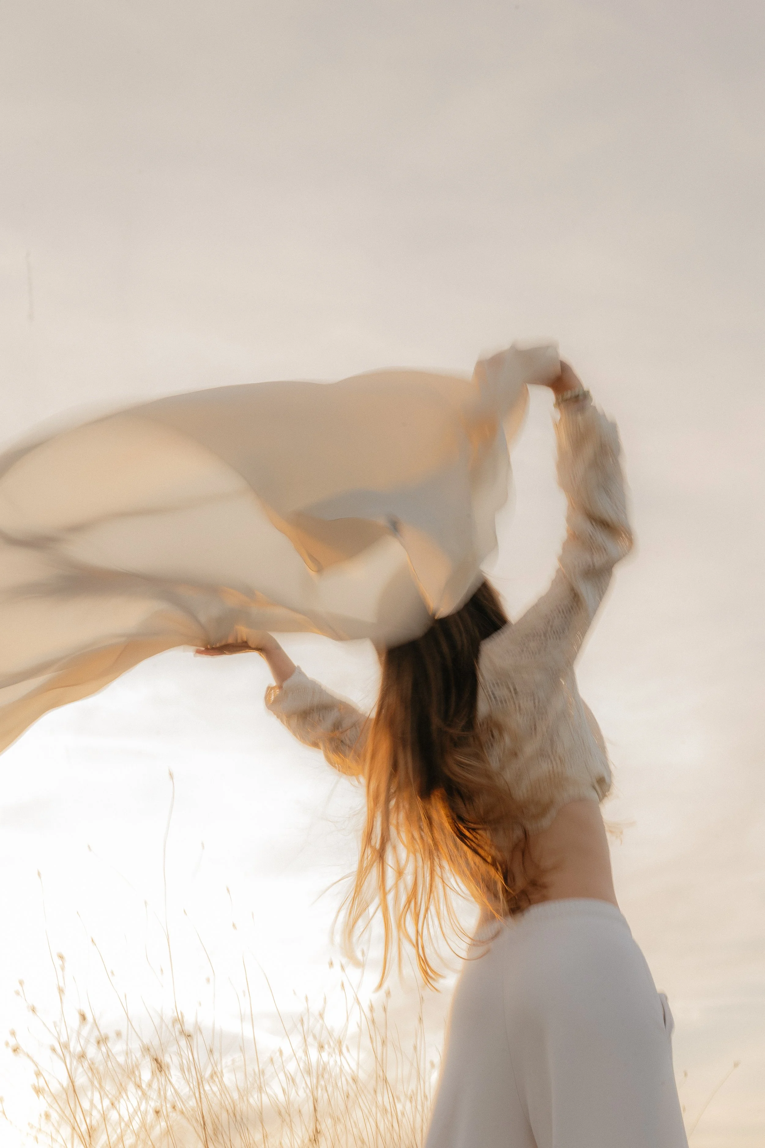 A woman with long hair stands outdoors, lifting a flowing piece of fabric in the air during sunset.
