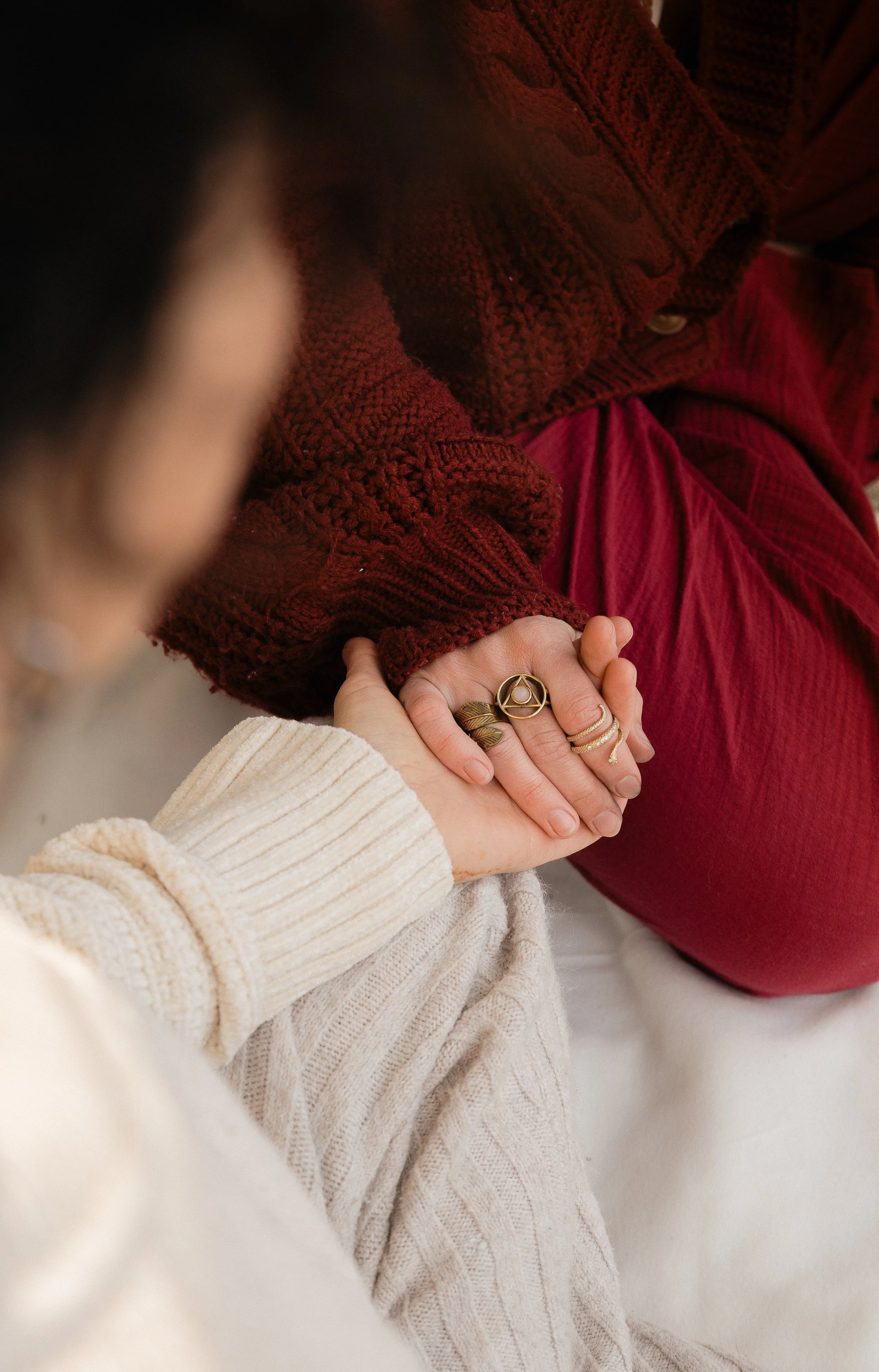 Two people holding hands, one with multiple rings on fingers, the other wearing a cream-colored, ribbed sweater, and the background person wearing a red sweater.