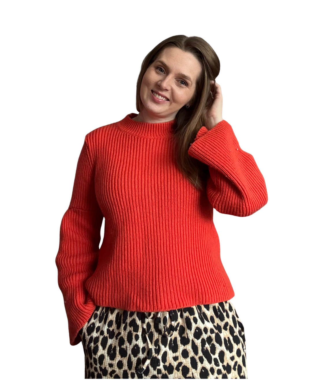A woman with brown hair wearing a red ribbed sweater and leopard print pants, smiling and touching her hair.