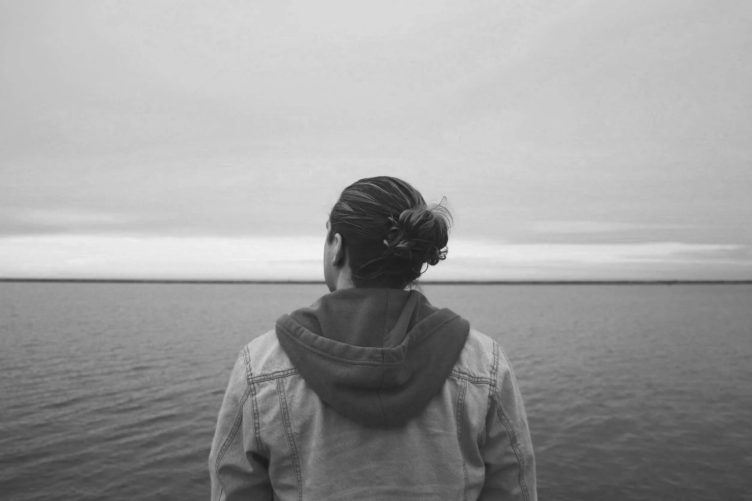 A person wearing a denim jacket and hoodie looking out over a calm body of water with a cloudy sky.