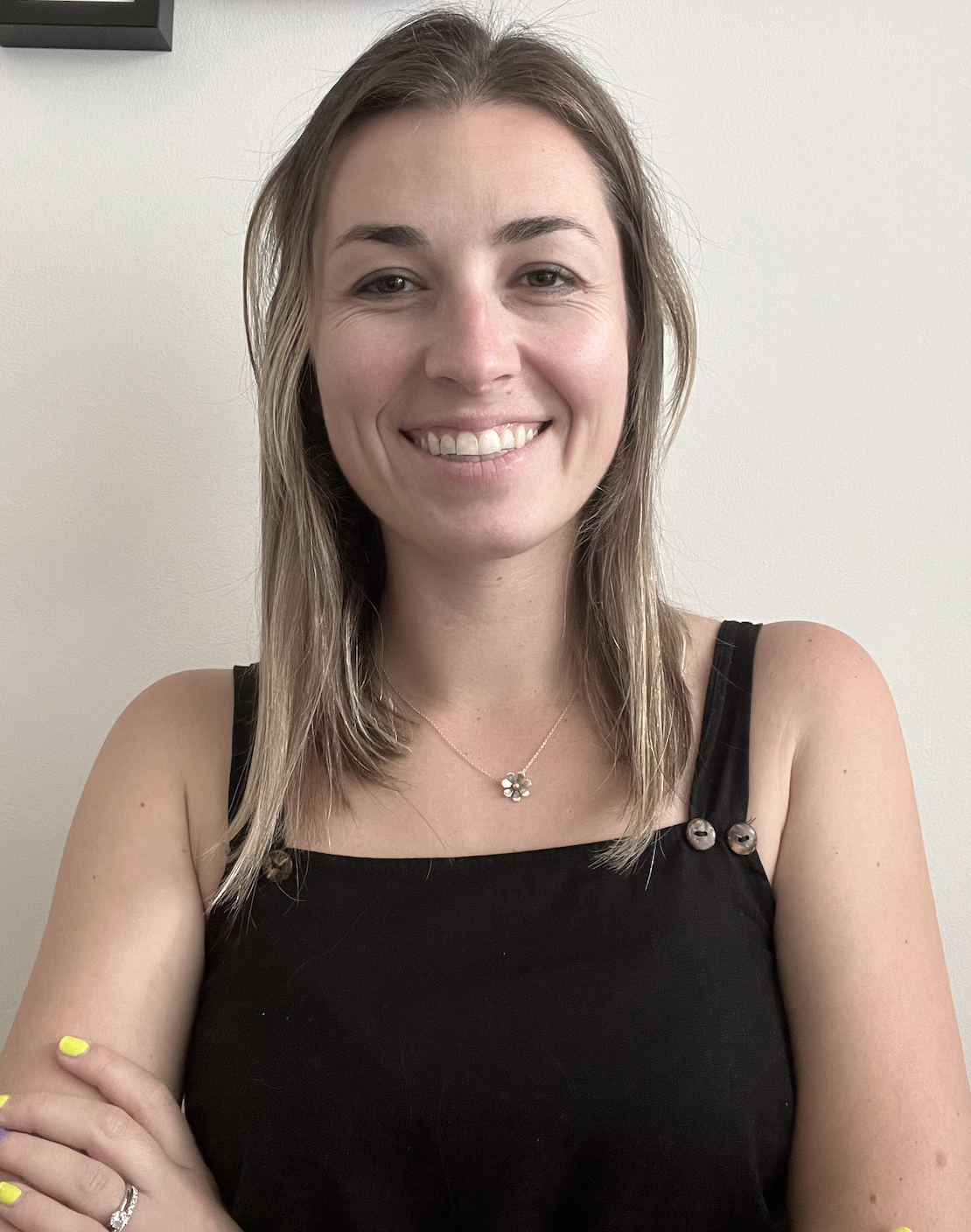 A woman with shoulder-length light brown hair, wearing a black sleeveless top with buttons on the straps, a silver necklace with a flower-shaped pendant, and a wedding ring, stands against a plain off-white wall and smiles at the camera.