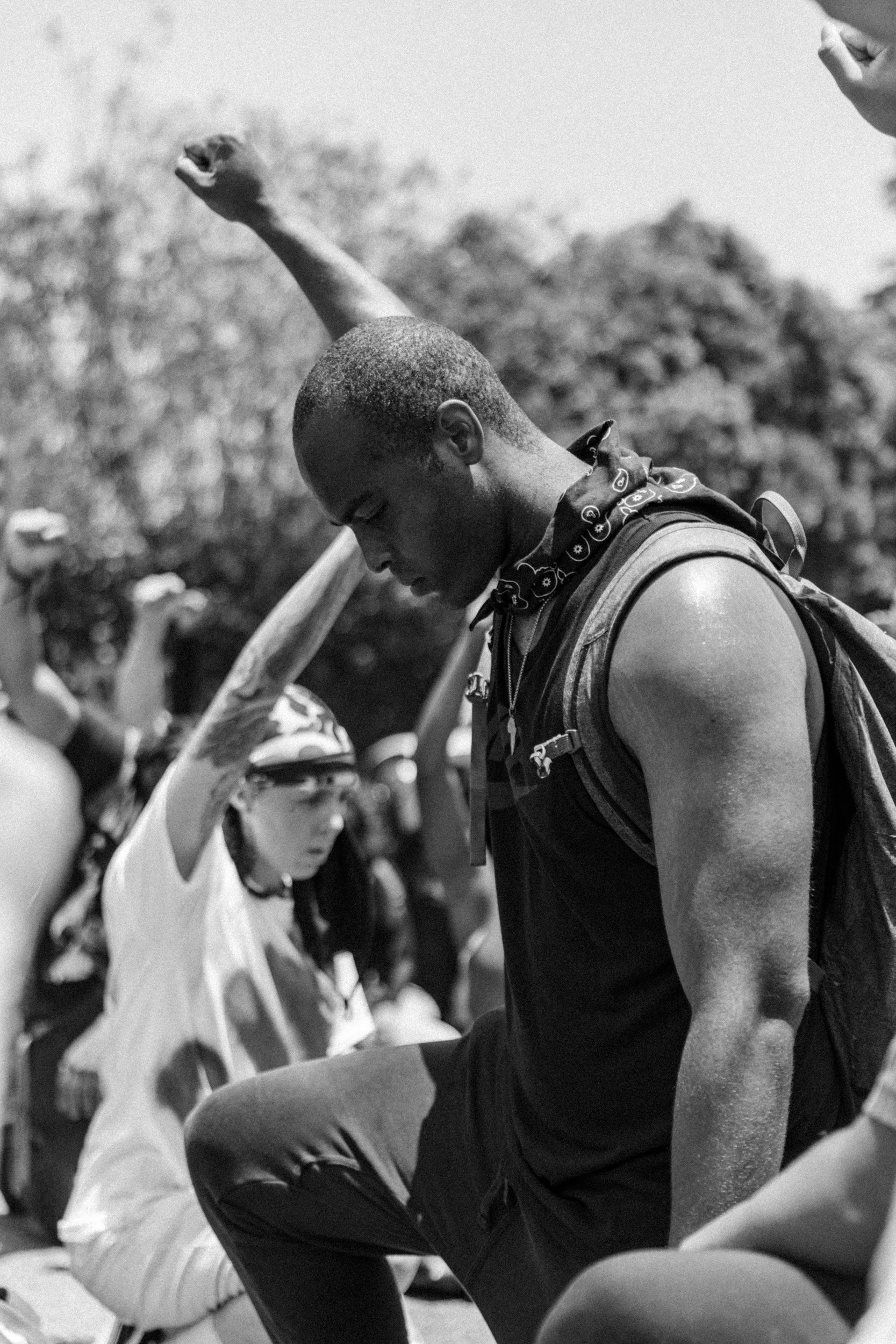Young man in athletic attire with a backpack, head bowed, and a girl with sunglasses and a bandana raising her fist in a crowd outdoors.