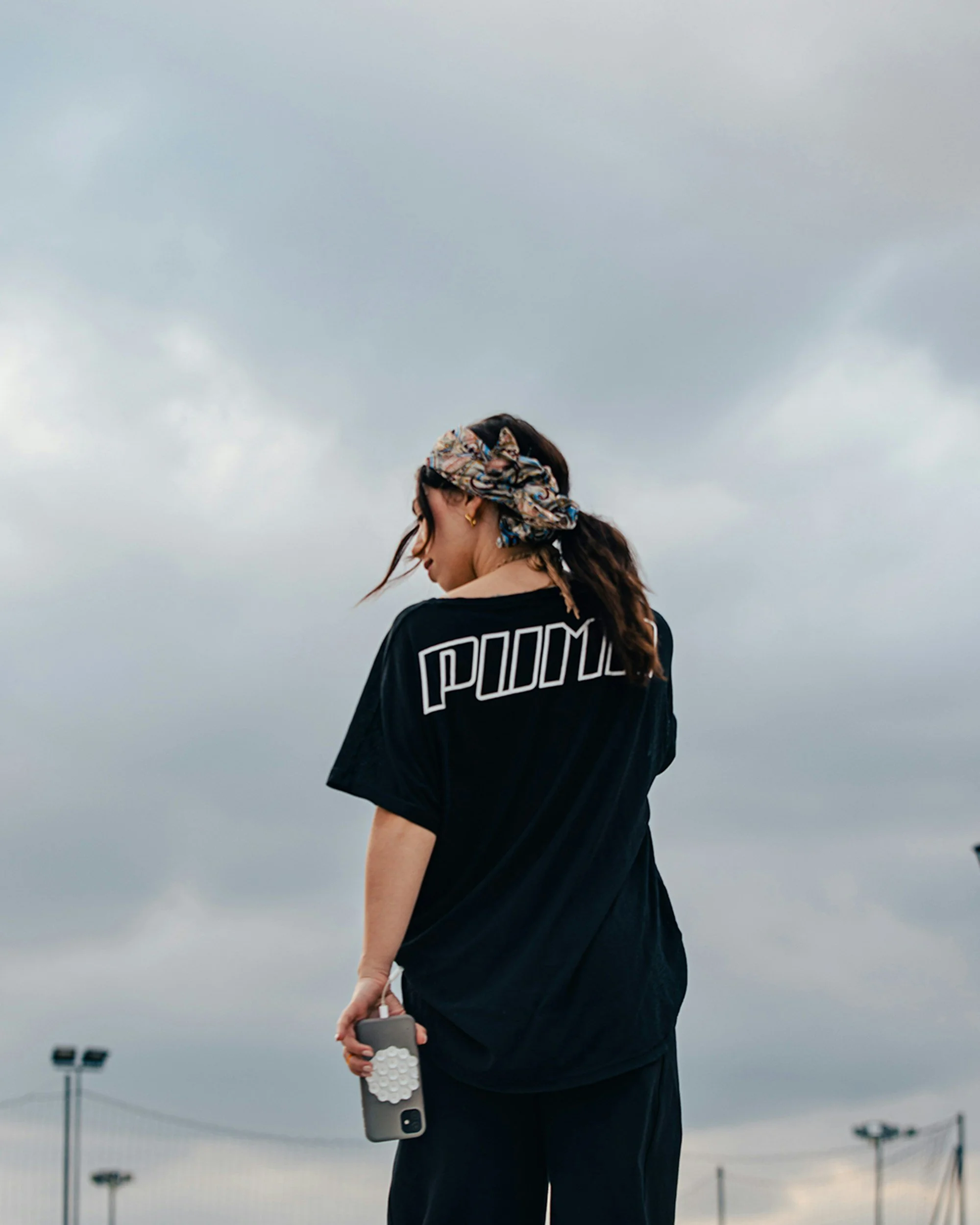 A woman with a patterned headscarf and earrings, wearing a black oversized sports jersey, holding a cell phone with a honeycomb-patterned case, standing outdoors under a cloudy sky.