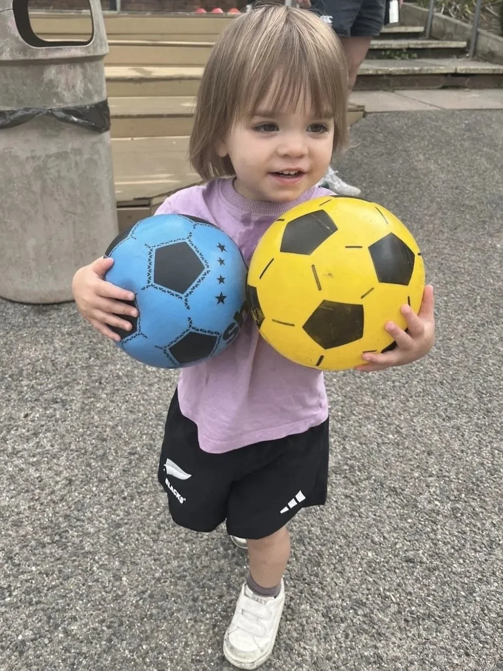 Young child holding two soccer balls, one blue and one yellow, outdoors near a wooden bench.