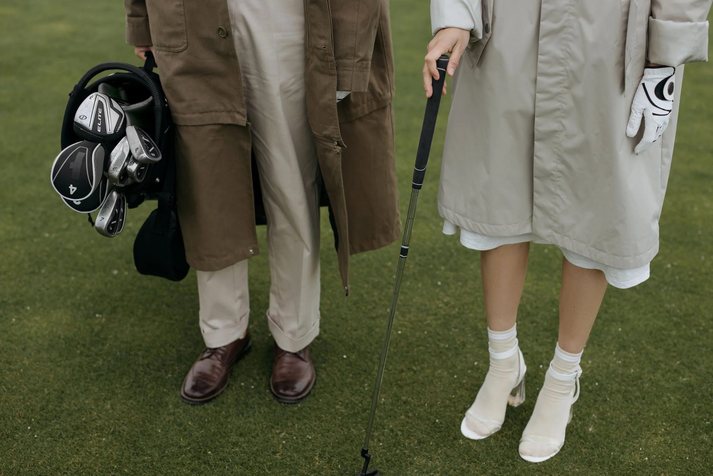 Close-up of two people standing on a golf course, showing their legs and golf clubs, with one person holding a golf club and the other person holding a golf glove.