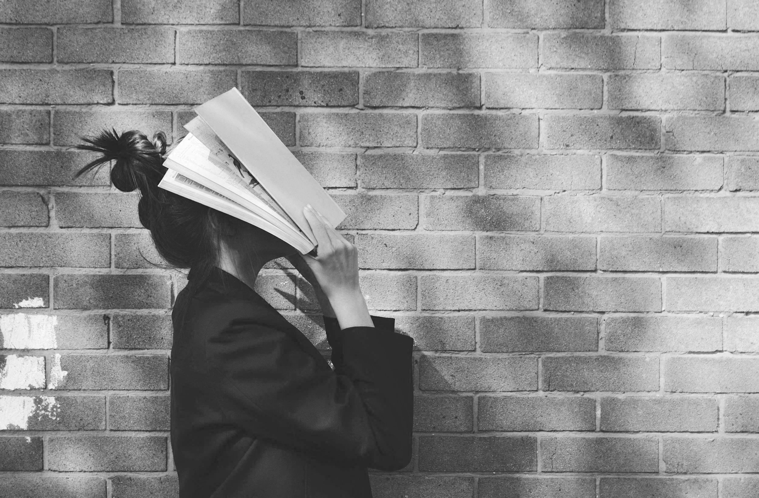 A person with their hair in a bun, wearing a dark jacket, is holding a magazine up to their face against a brick wall.