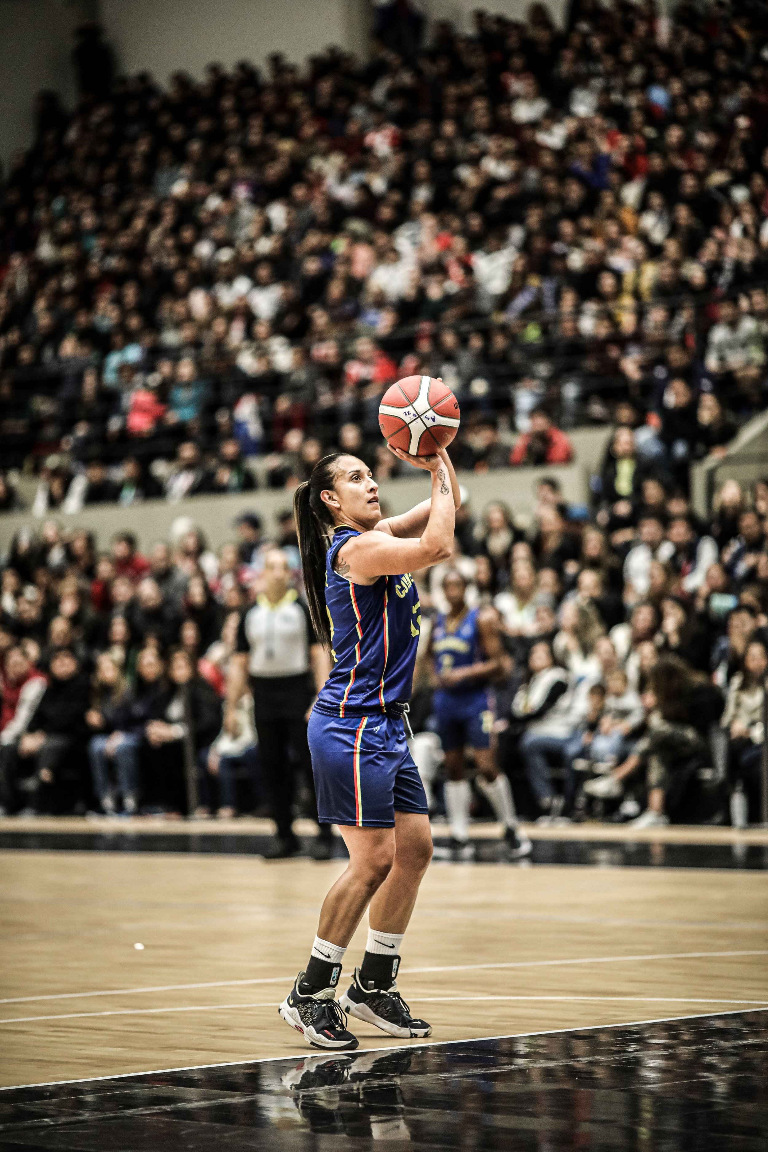 A female basketball player in a blue uniform holding a basketball on a basketball court with a large crowd of spectators in the background.