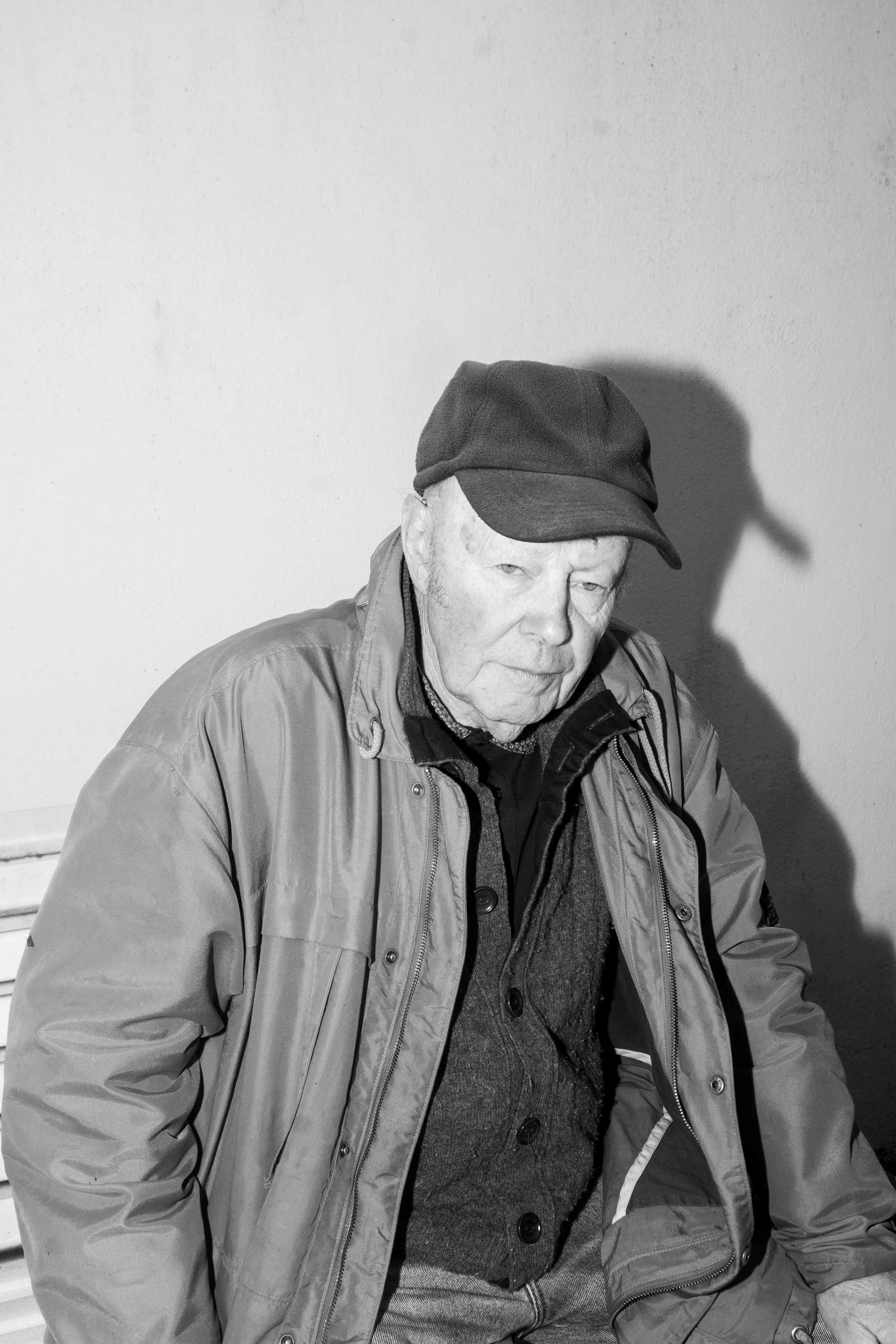 Un vieil homme portant une casquette et un manteau, assis et regardant vers le bas, dans une pièce avec un mur blanc.