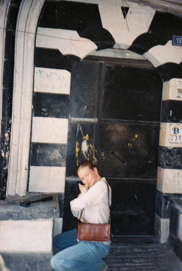 A young woman sitting on a bench hugging a small dog in front of a black and white striped door with an arched top, in an urban setting. Emilysfamous syria tour