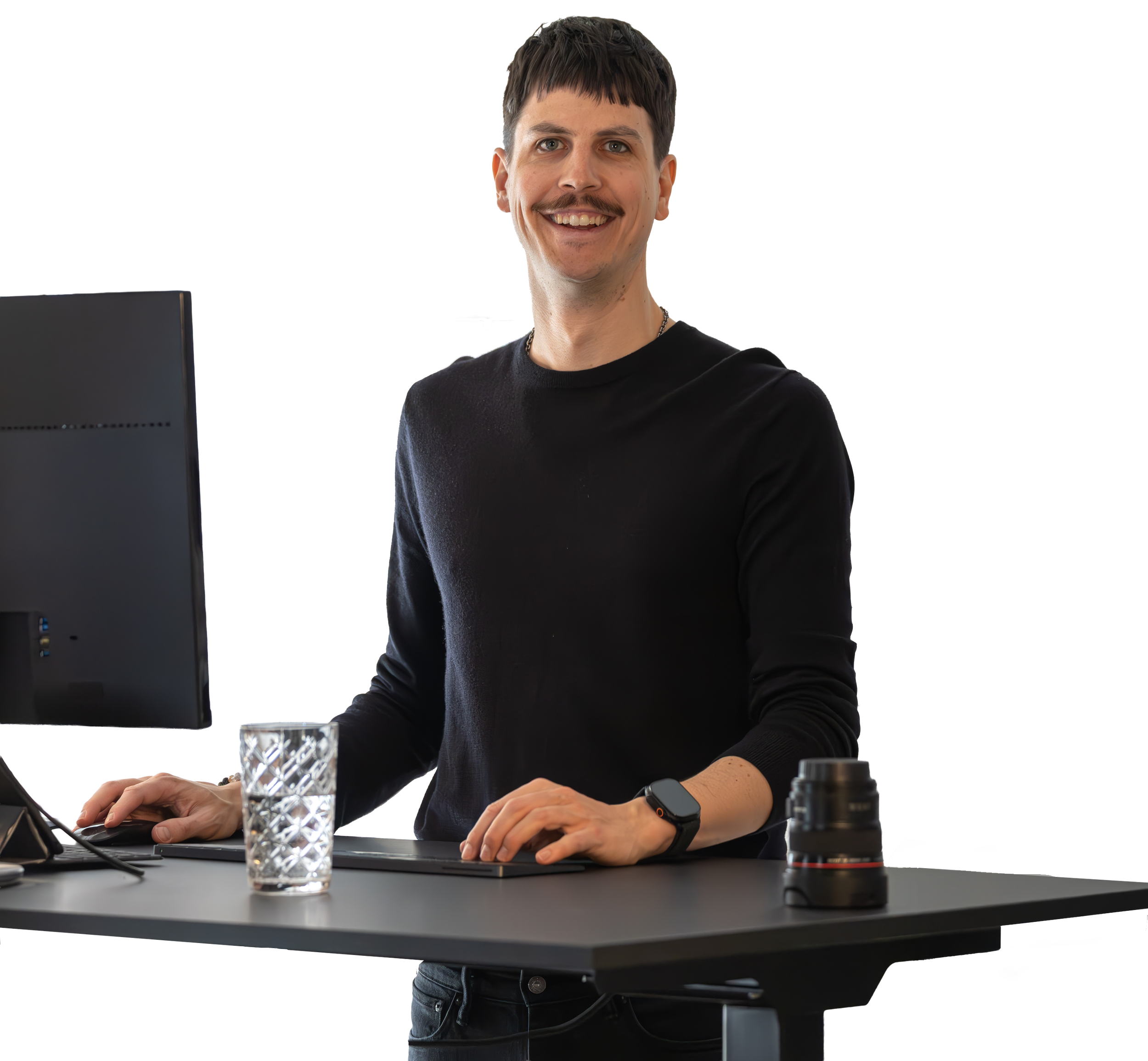Mann steht an einem Schreibtisch mit Monitor, Glas Wasser, Kamera und Tastatur, trägt schwarze Kleidung, Smartwatch, lächelt, dunkle Haare, kurzen Haarschnitt.