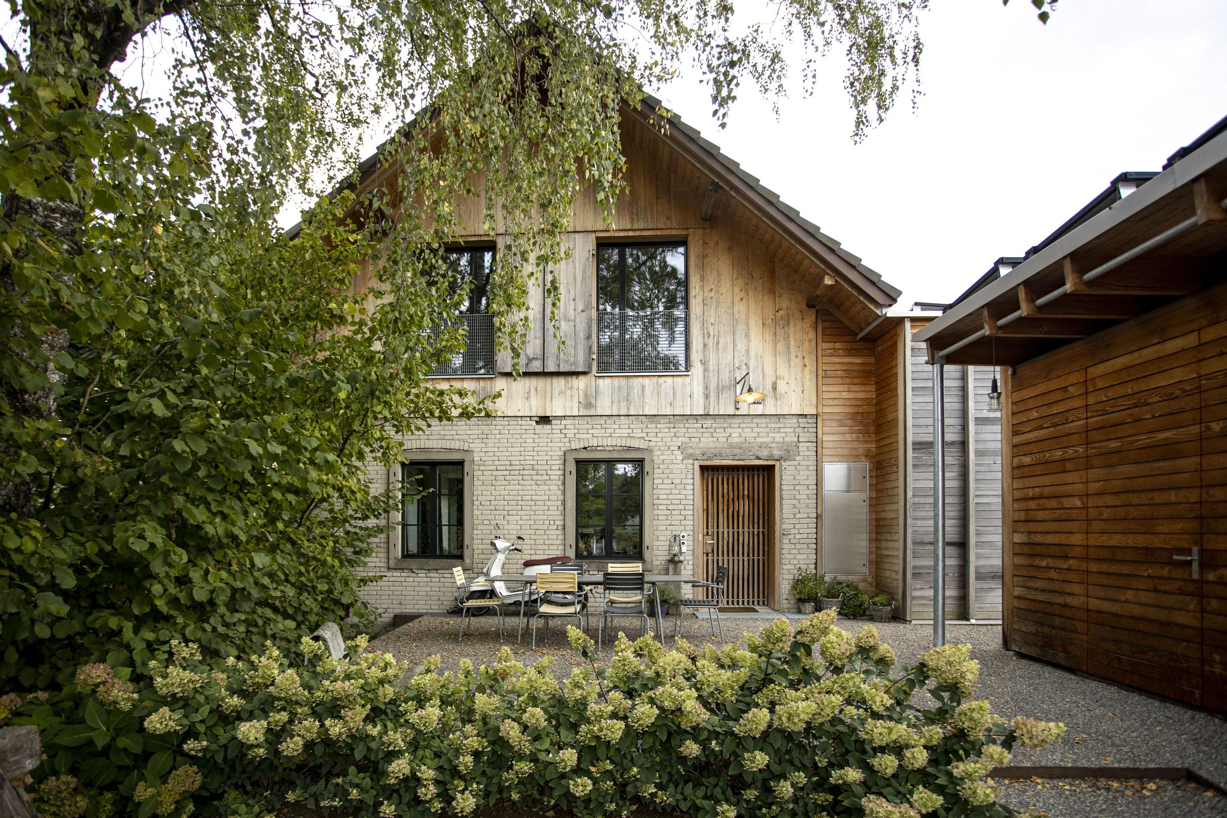 Ein modernes, zweistöckiges Haus mit gemischtem Außenmaterial aus Holz und Ziegel, umgeben von grünen Pflanzen und einer Terrasse mit Möbeln im Garten.